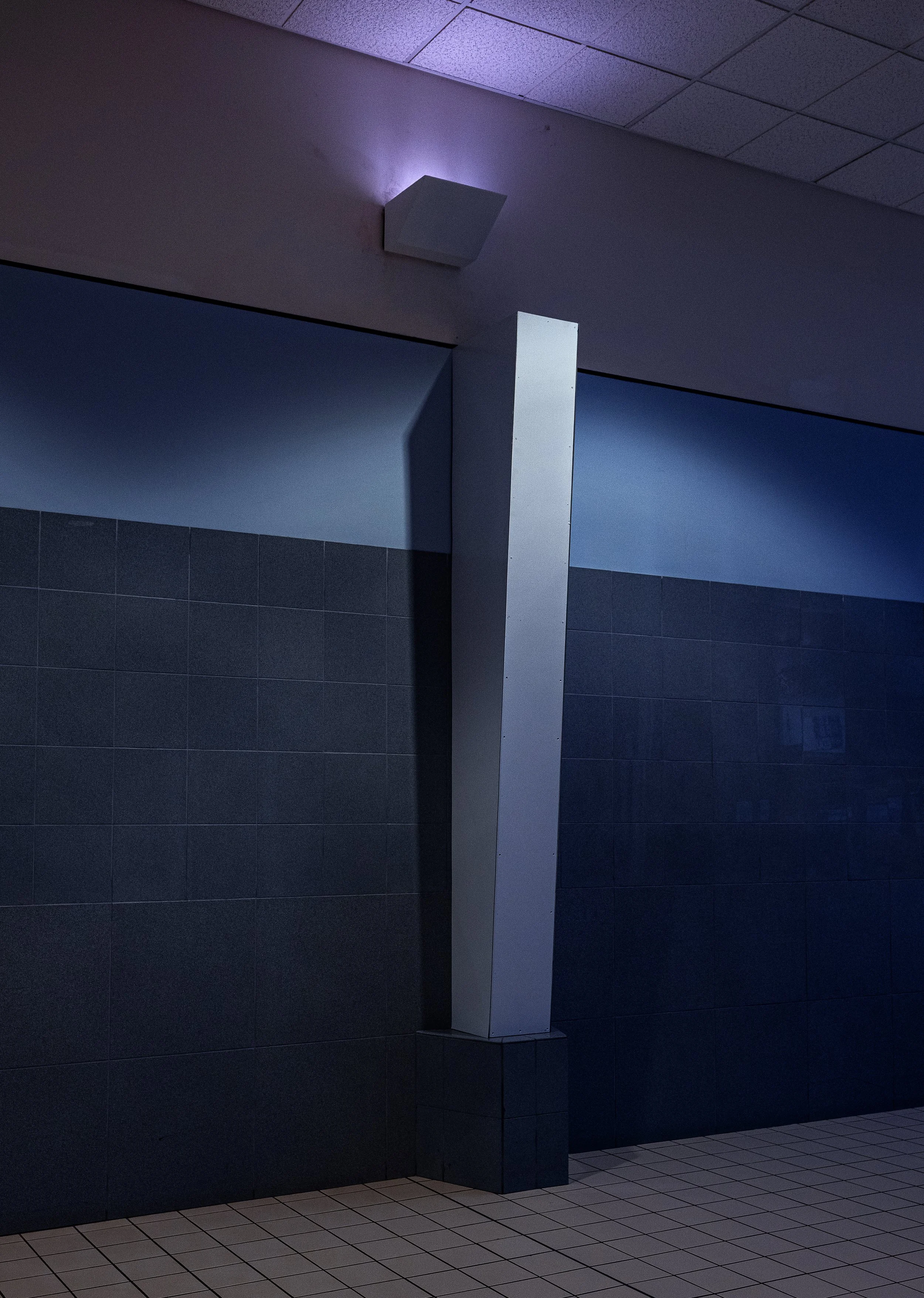 A white rectangular column stands next to a dark tiled wall in a dimly lit indoor environment, with a ceiling mounted cube-shaped light fixture emitting purple light, against a background of dark tiles and a tiled floor.