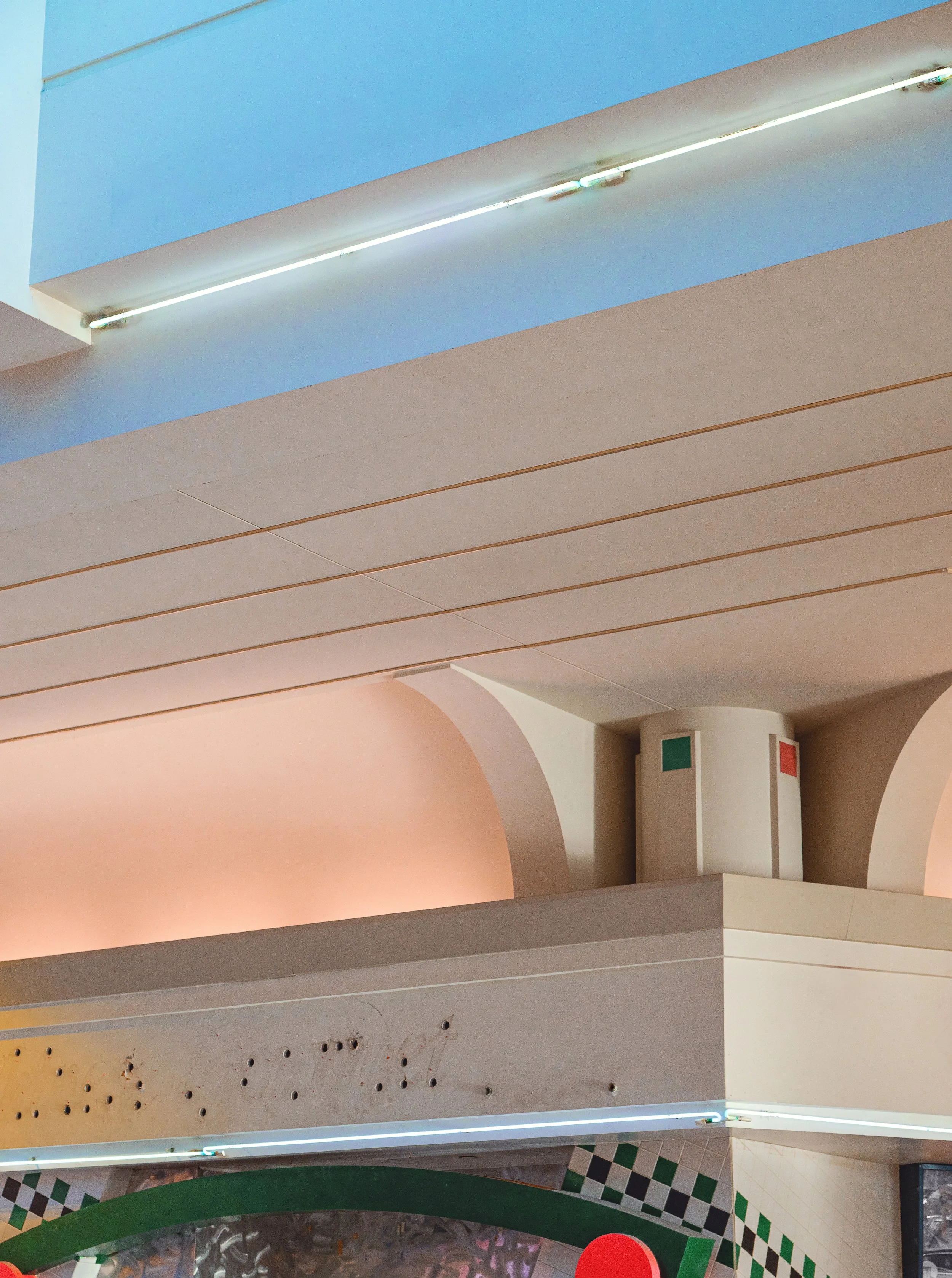 Indoor modern architectural ceiling with curved and flat surfaces in earth tones, illuminated by linear lighting fixtures, with a column featuring a flag motif and a checkered pattern on the walls and floors.