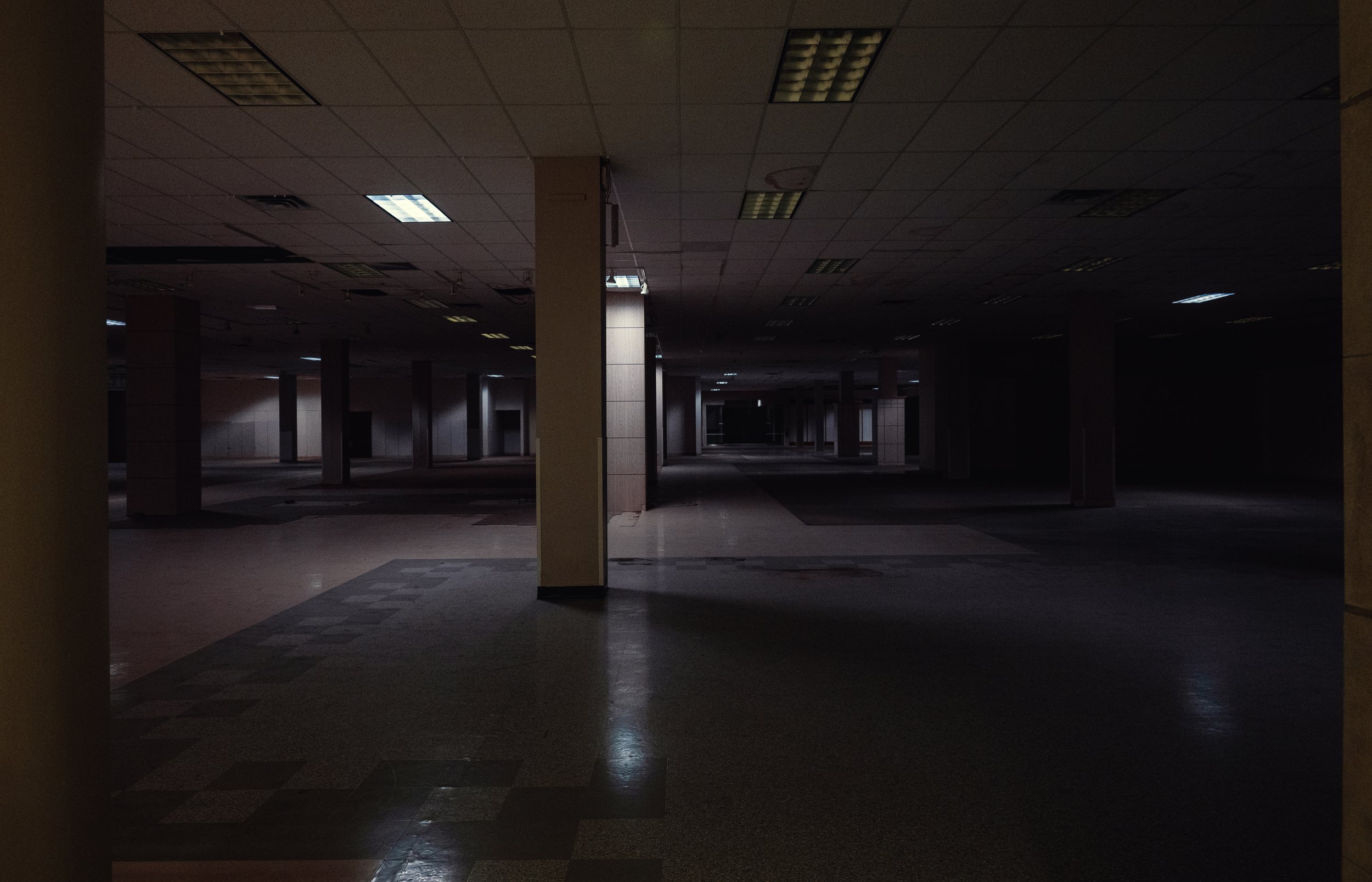 A dark, empty indoor space with a tiled ceiling and fluorescent lights, supported by several columns, with some light visible in the distance.