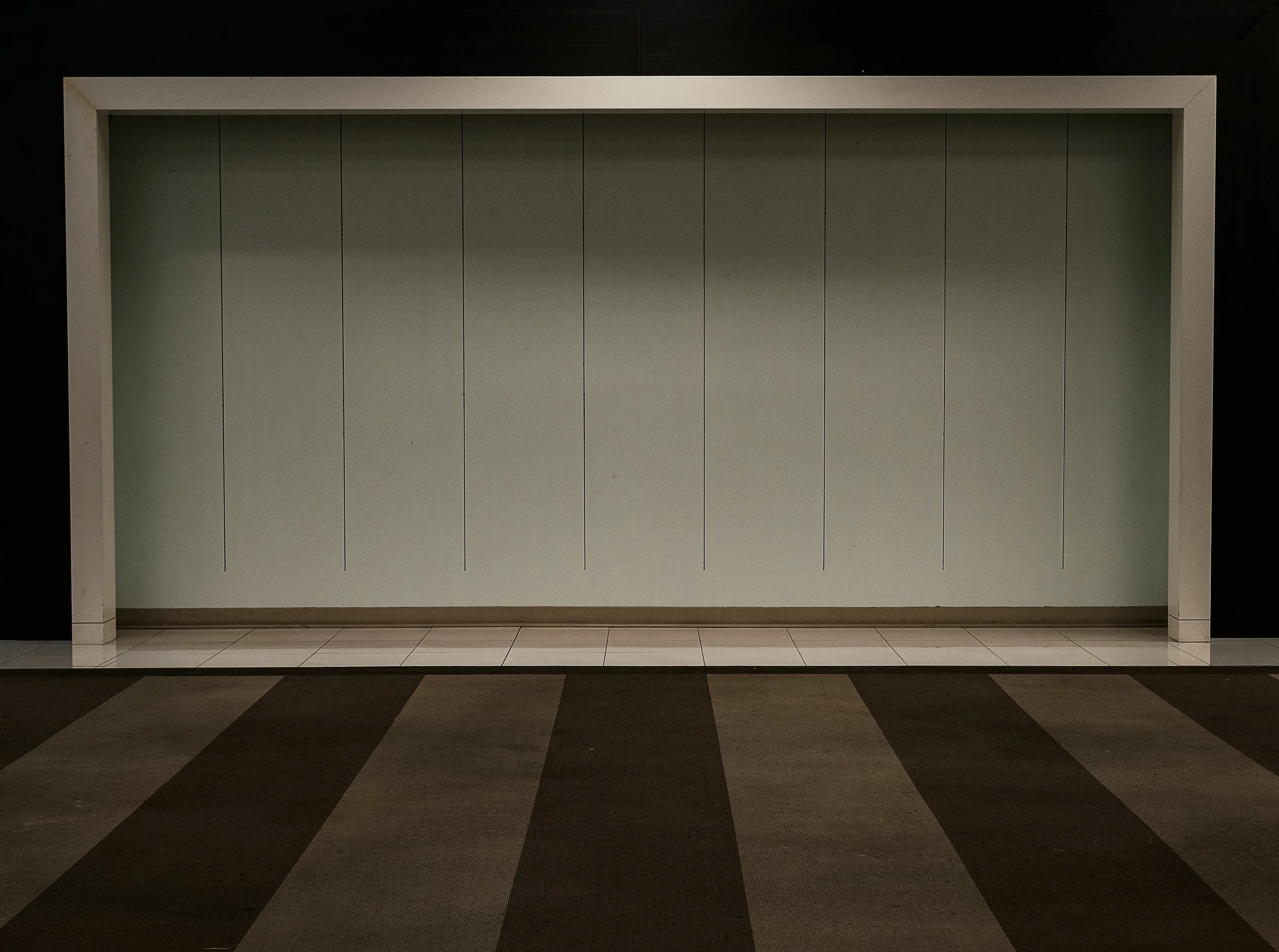 Empty retail or exhibition space with a plain light green wall, white trim, and checkered carpeted floor.