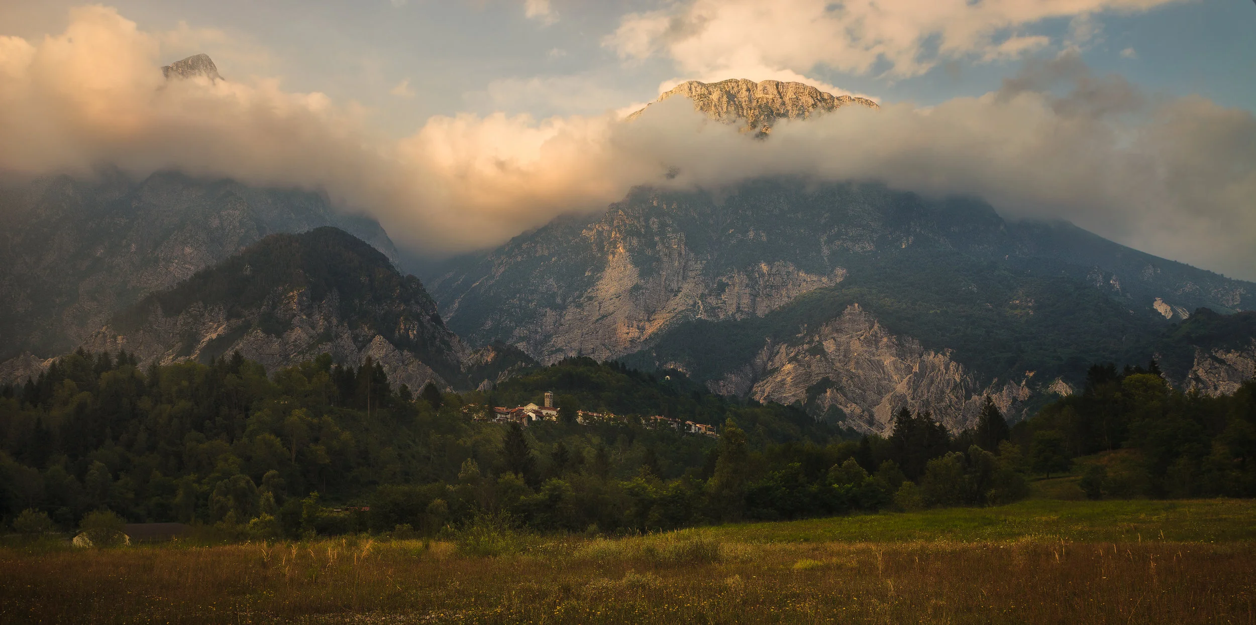 Mountain peaks covered in clouds at sunset at Barcis Lake, Italy. The face of the mountains above a quaint mountainside town peaks through the clouds and is lit by the warm sunlight.