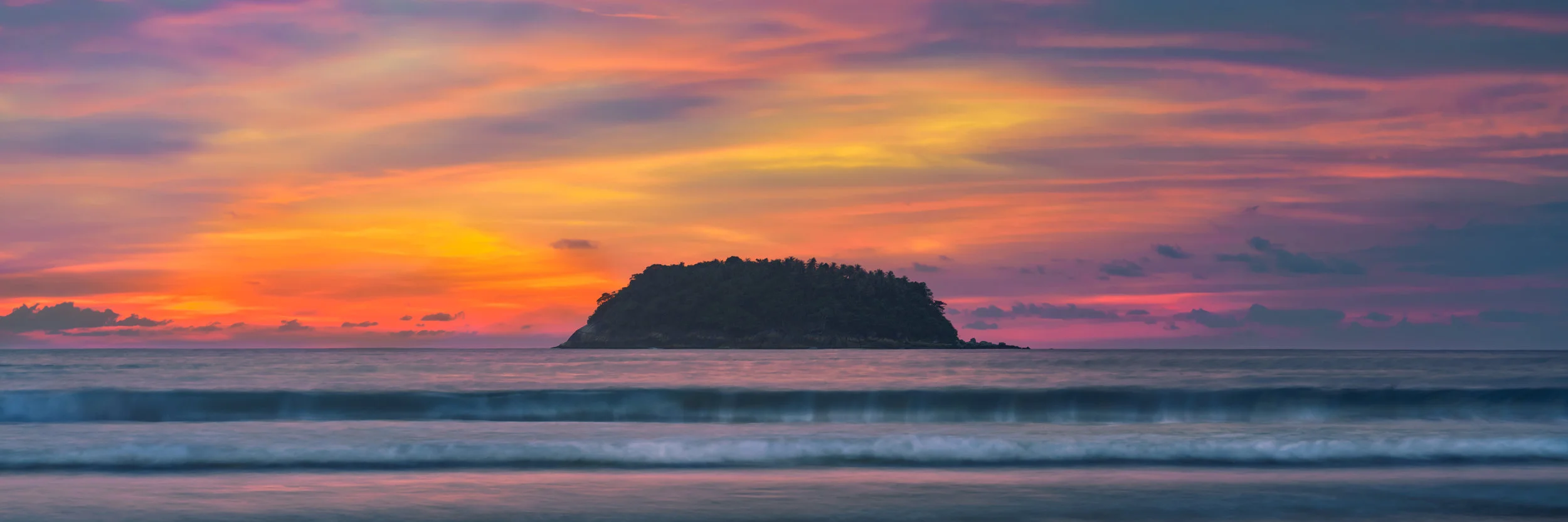 There aren't many words to describe a phenomenal sunset. When you say you've seen one the person with you pictures it in their mind's eye. For me to say, this night was phenomenal would do little justice to just how astounded I was that the sky could make some many colors at once. Phuket blessed us with two such nights. I captured one. I hunted for a clear space perpendicular to the waves, took a knee, waited, and was rewarded with light like I have experienced few times. When the sets began pounding the surf I knew I had my shot. Kata is a sunset set photograph captured at Kata Beach in Phuket Sunset. Pinks, magentas, oranges, and yellows decorate the evening sky in a magical show of color while long sets of waves roll onto shore. Ko Pu island sits in the center of the photograph silhouetted against a colorful sky.