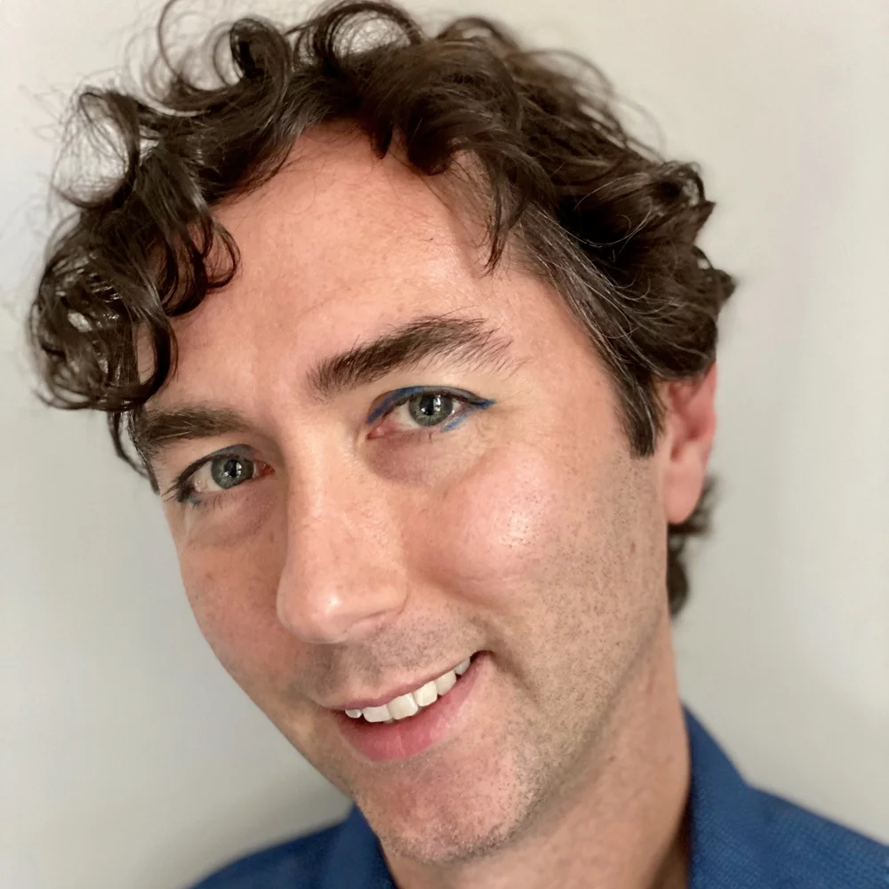 Headshot of Brian Gresko. Brian has light skin and short, curly brown hair and is smiling at the viewer with their head tilted toward the viewer's left. They are wearing a blue shirt and standing against a light gray wall.