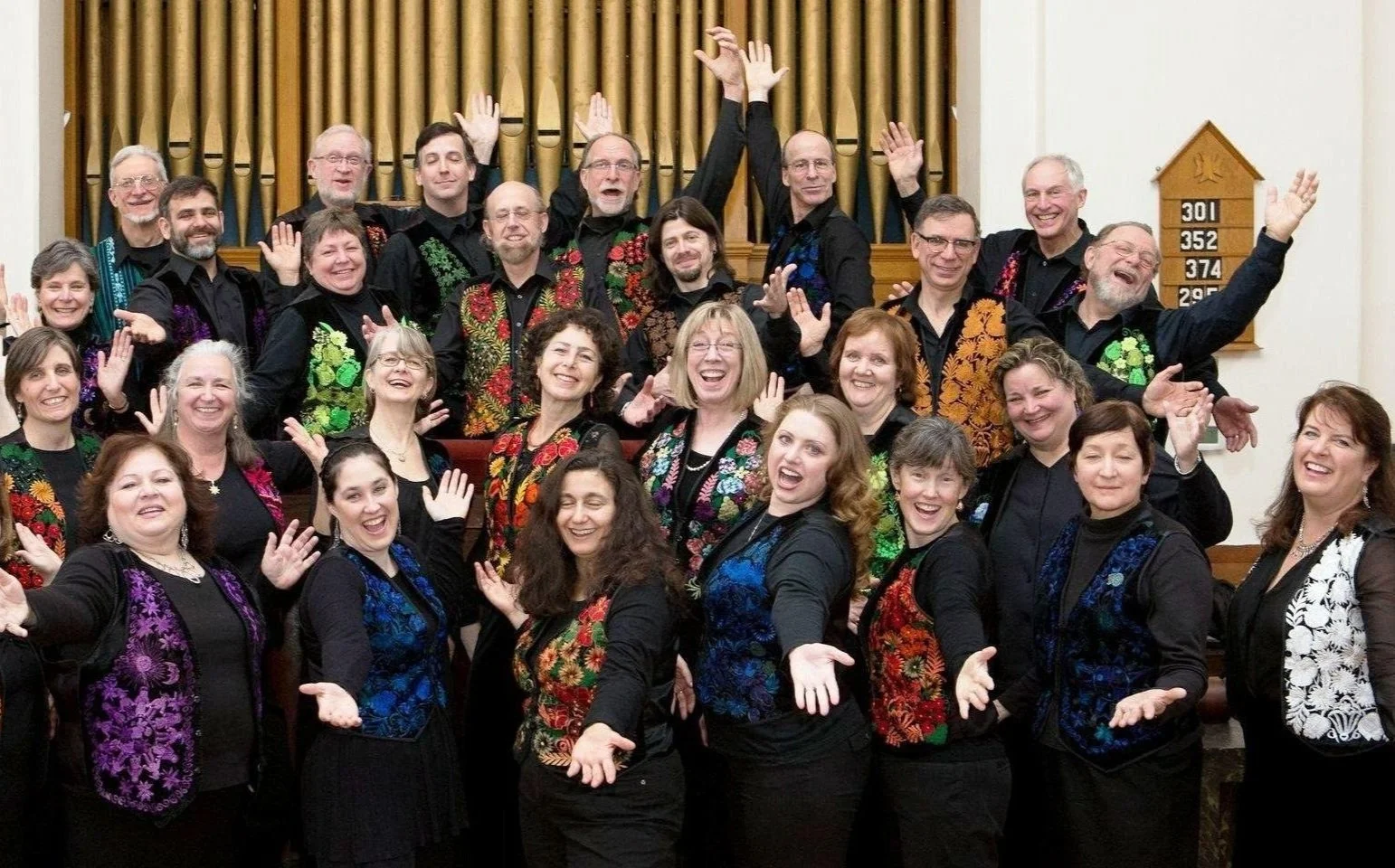 A large group of people dressed in colorful, embroidered choir robes, smiling and posing for a photo inside a church with wooden pipes in the background.
