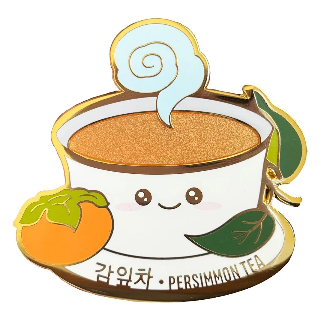 Cute enamel pin of tea in a white cup with a persimmon and leaves.