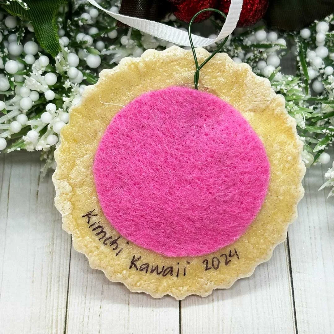 Back of a round Christmas cookie ornament with felt backing and a signature that says 'Kimchi Kawaii, 2024'.