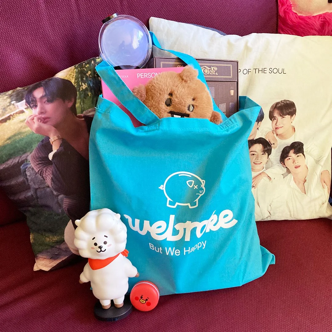 Various kpop items in a blue tote bag with white screen printed design of a sad piggy bank and the words, 'Webroke, but we happy'.