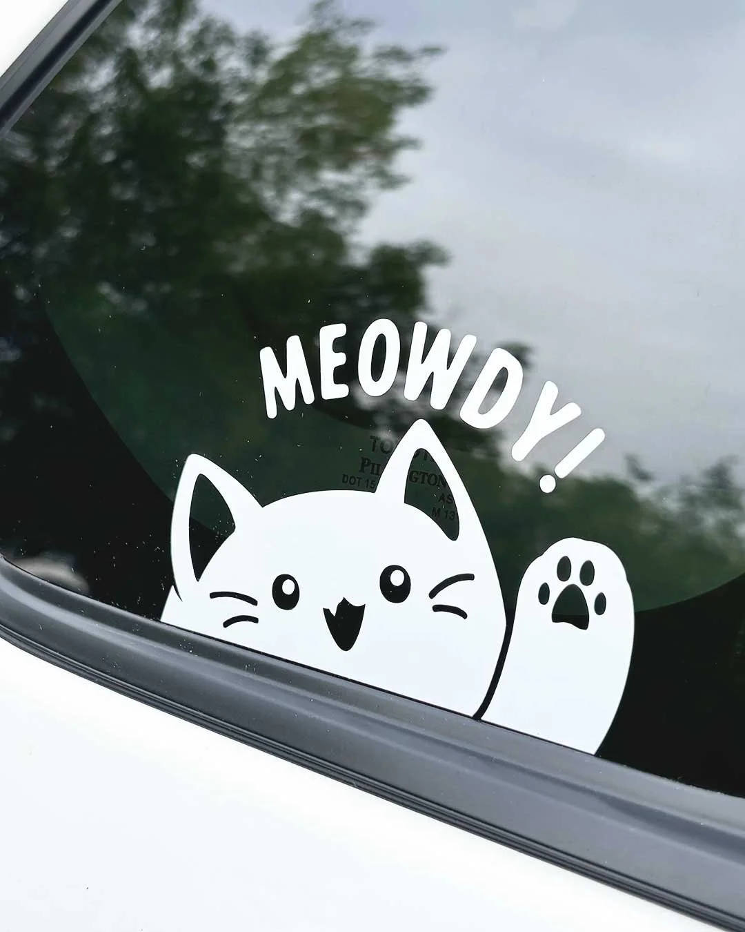 White vinyl sticker of a cartoon kitty waving with one paw and saying 'Meowdy'.