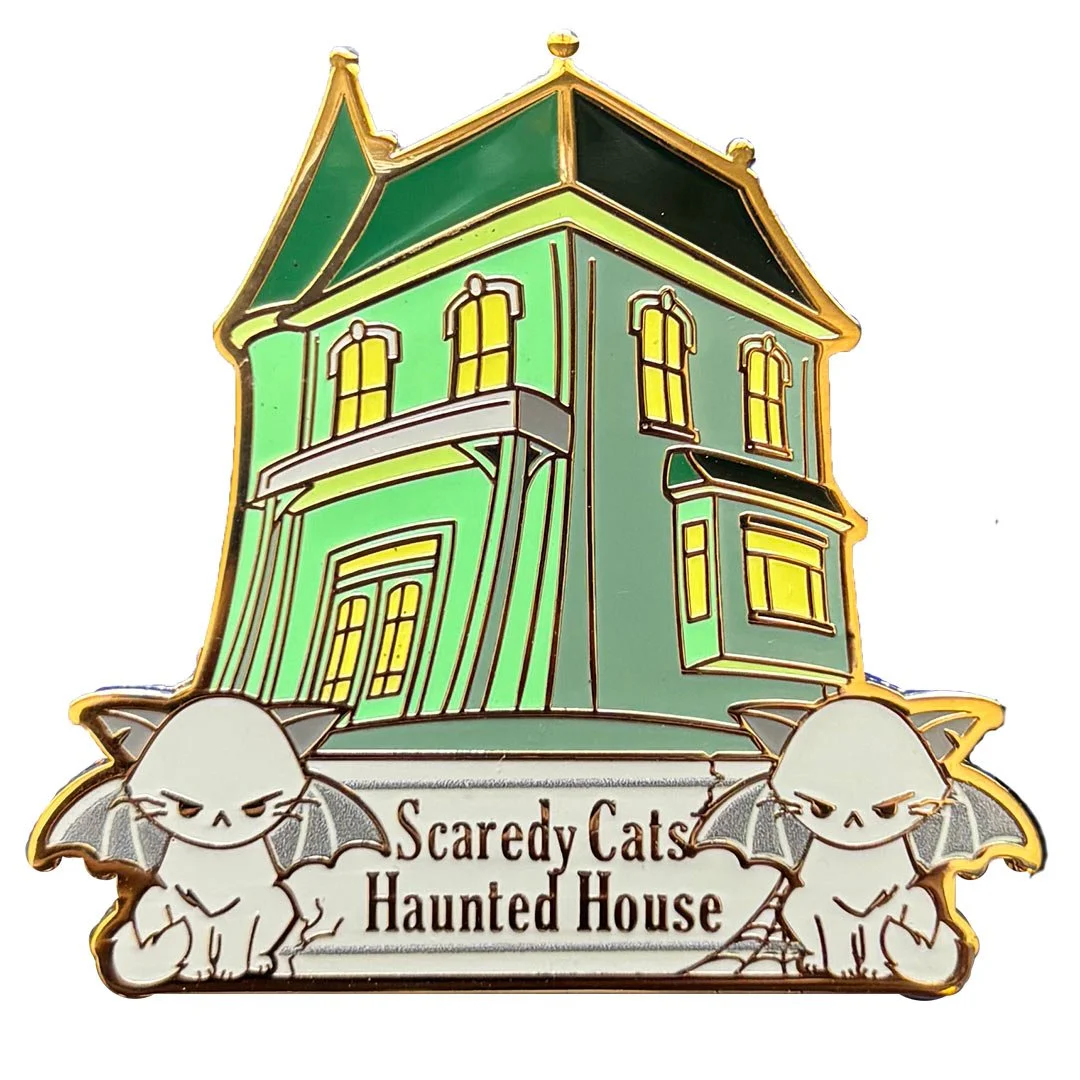 Enamel pin of a green haunted house with yellow windows and grey cat bats sitting on either side of a sign with the words 'Scaredy Cats Haunted House'.