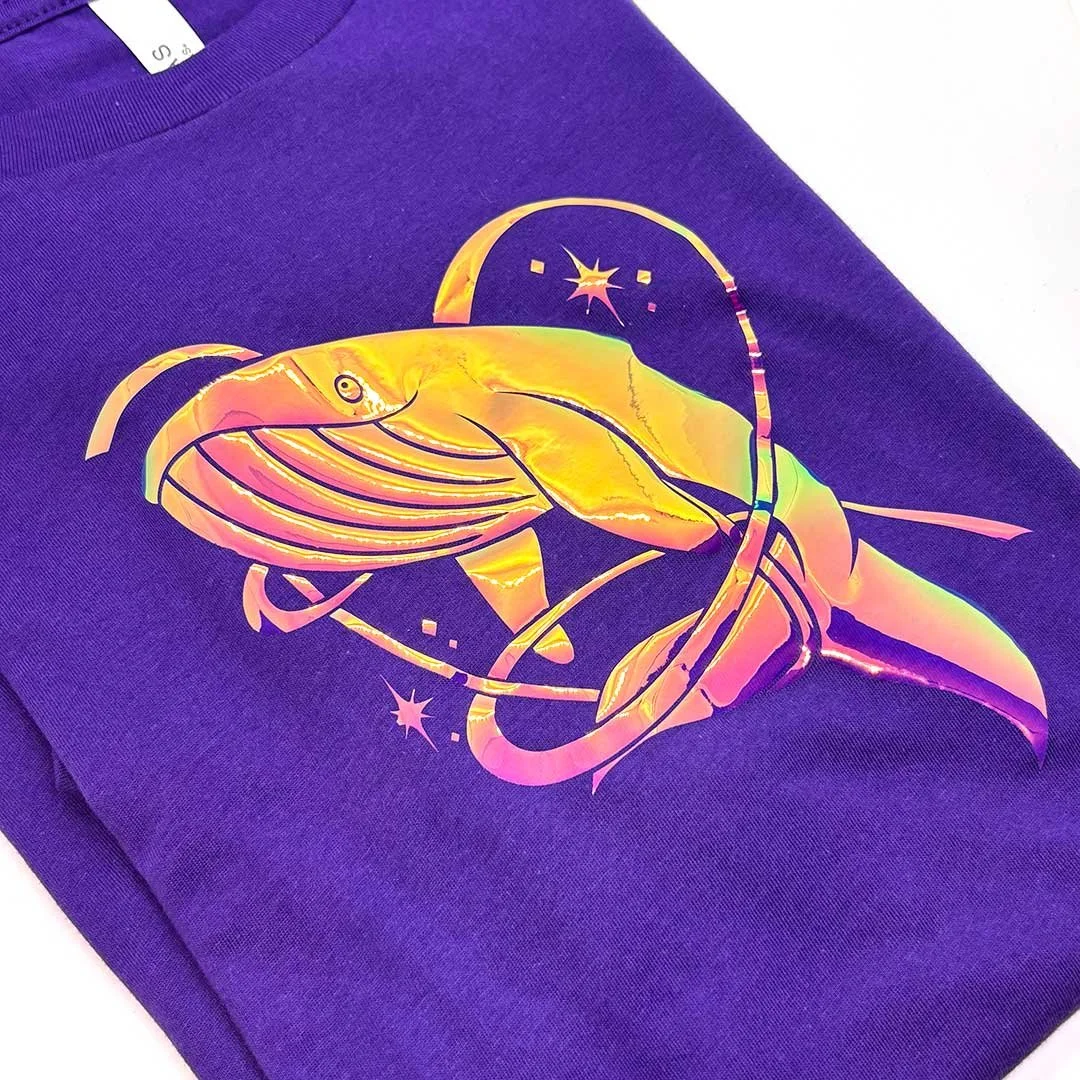 Humpback whale with a stylized heart around it in holographic iron on a purple shirt.