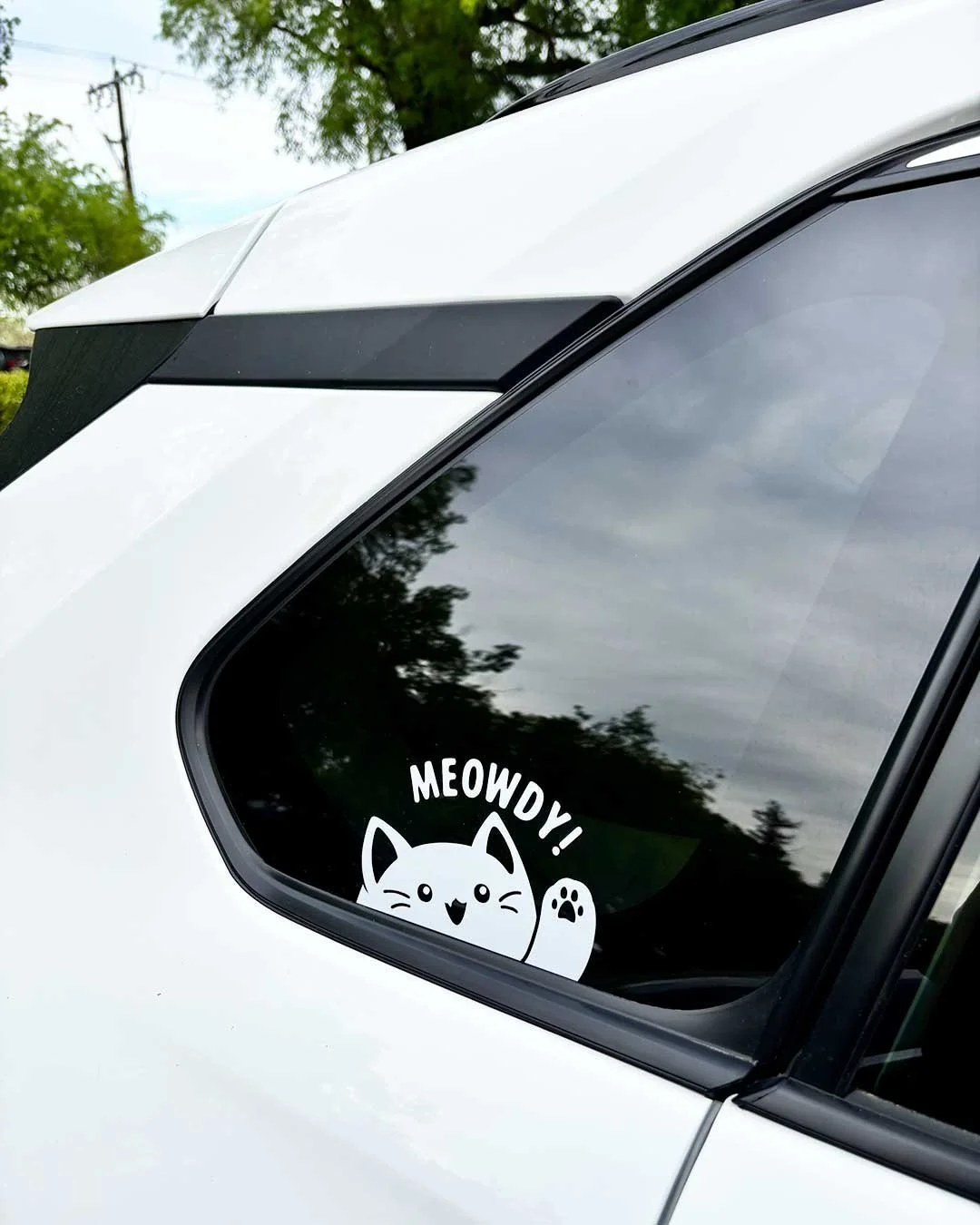 White vinyl sticker of a cartoon kitty waving with one paw and saying 'Meowdy'.
