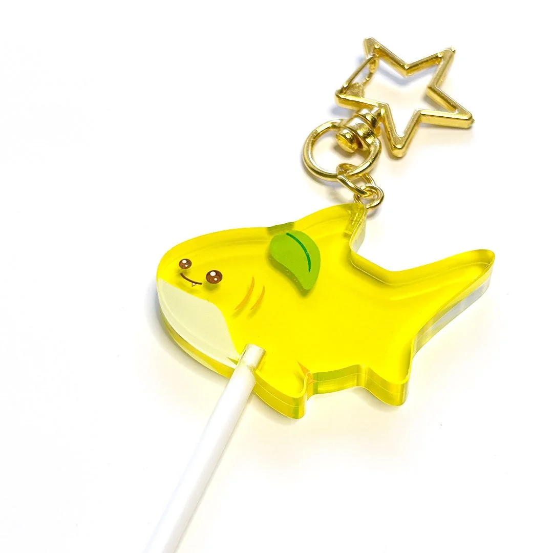 Detail of a yellow acrylic lemon shark lollipop charm with a gold clasp.