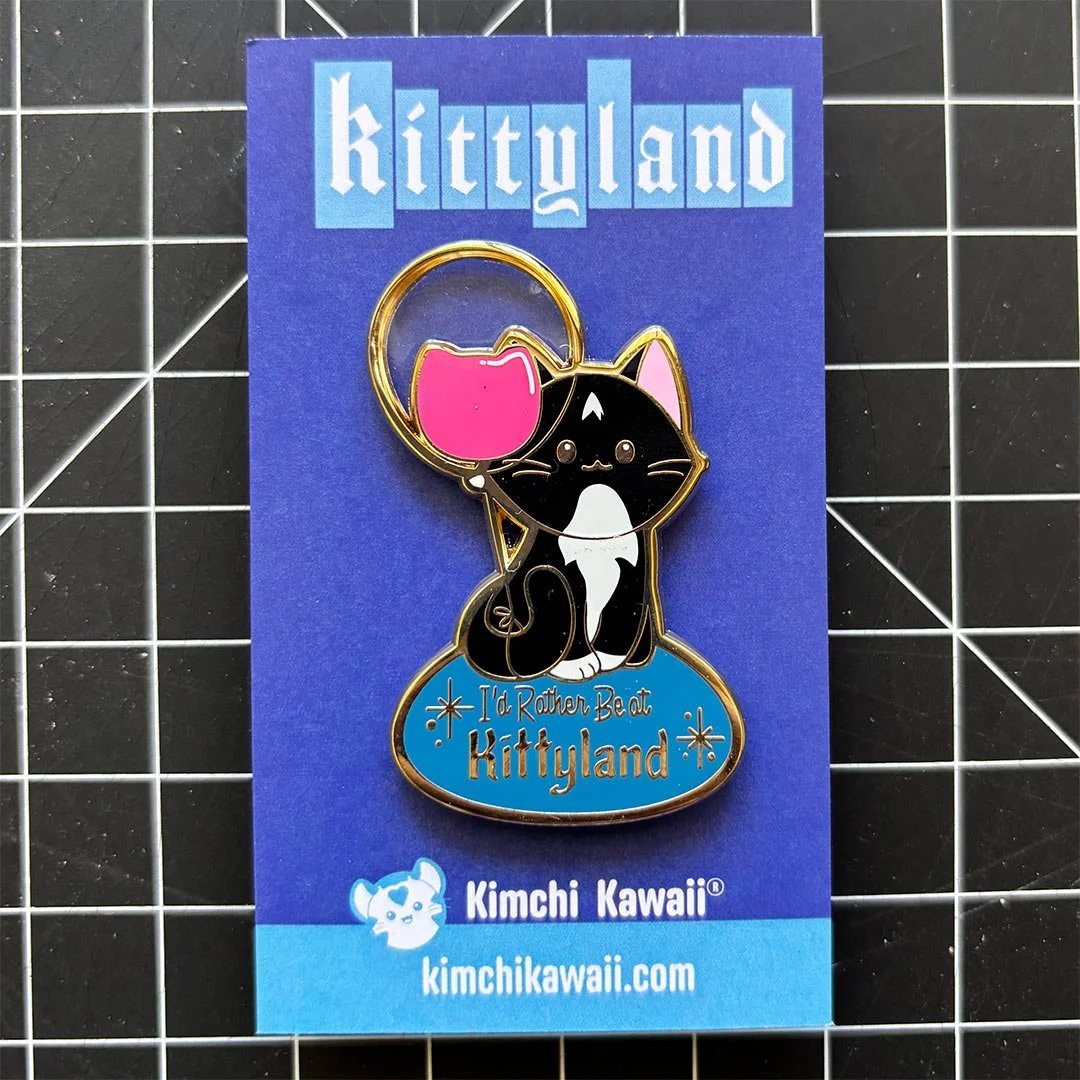 Enamel pin of a black and white kitty with a balloon. Text below says 'I'd rather be at Kittyland'.