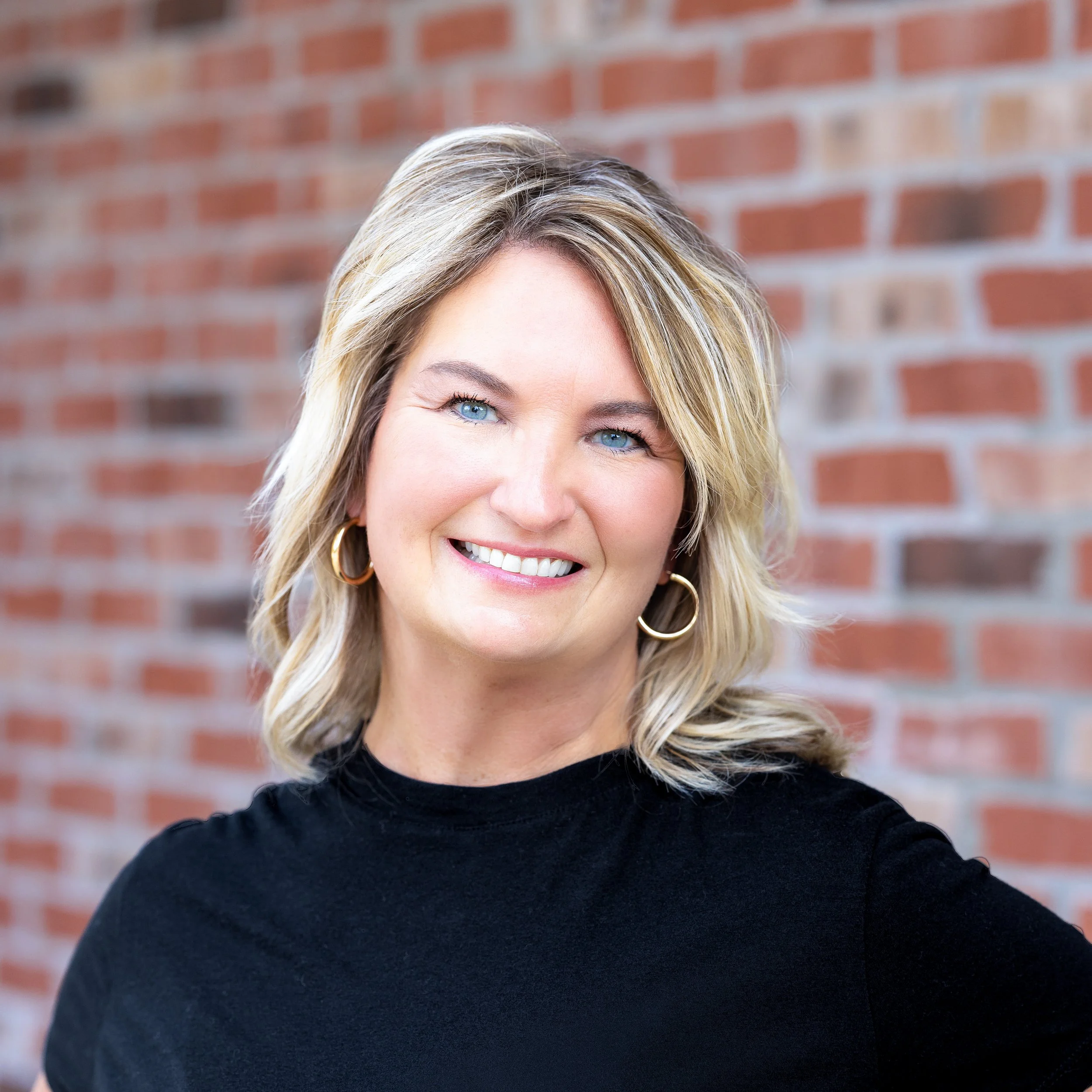 A woman with shoulder-length blonde hair, blue eyes, and earrings, smiling in front of a brick wall.