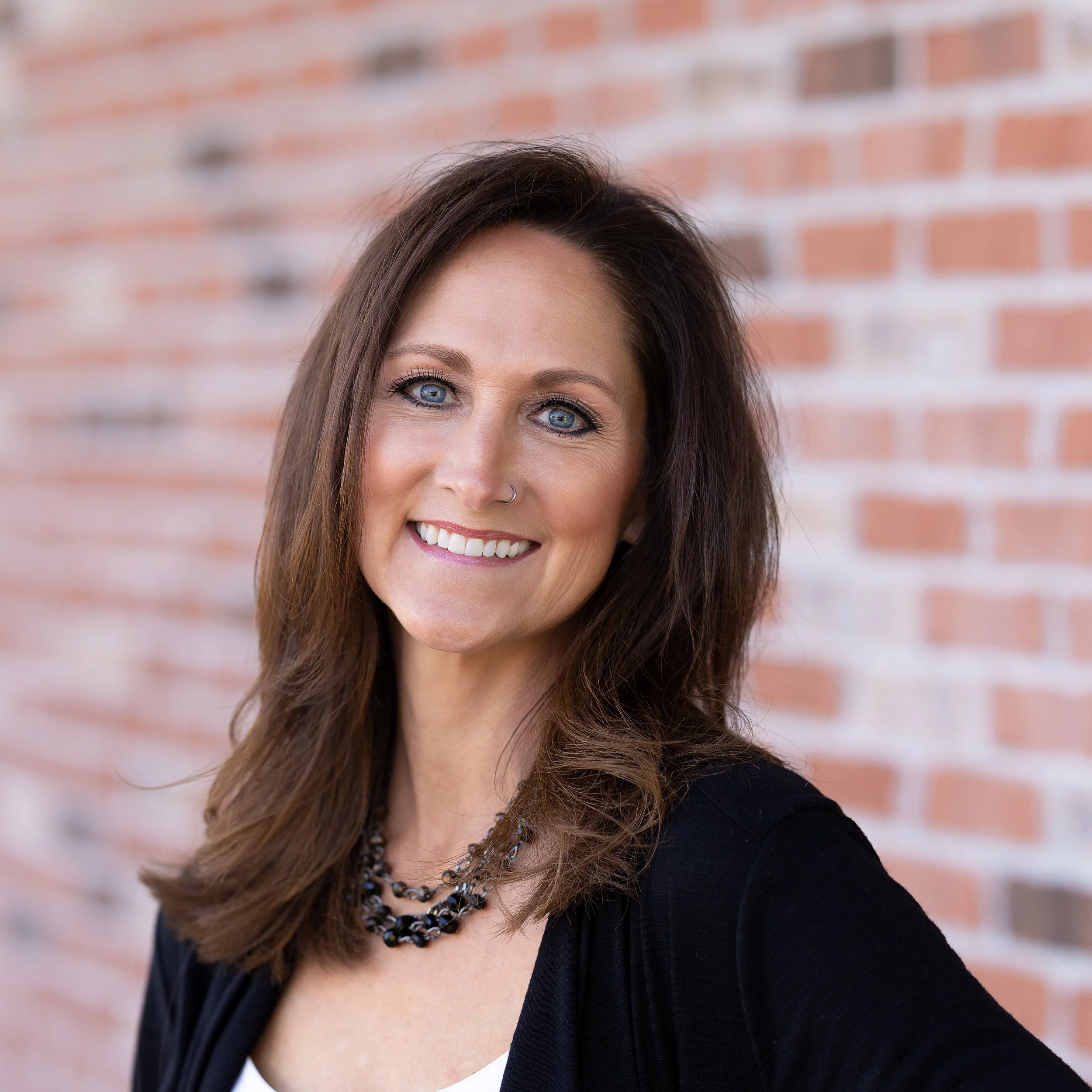 A smiling woman with brown hair, blue eyes, wearing a black jacket over a white top and a black beaded necklace, standing against a brick wall.