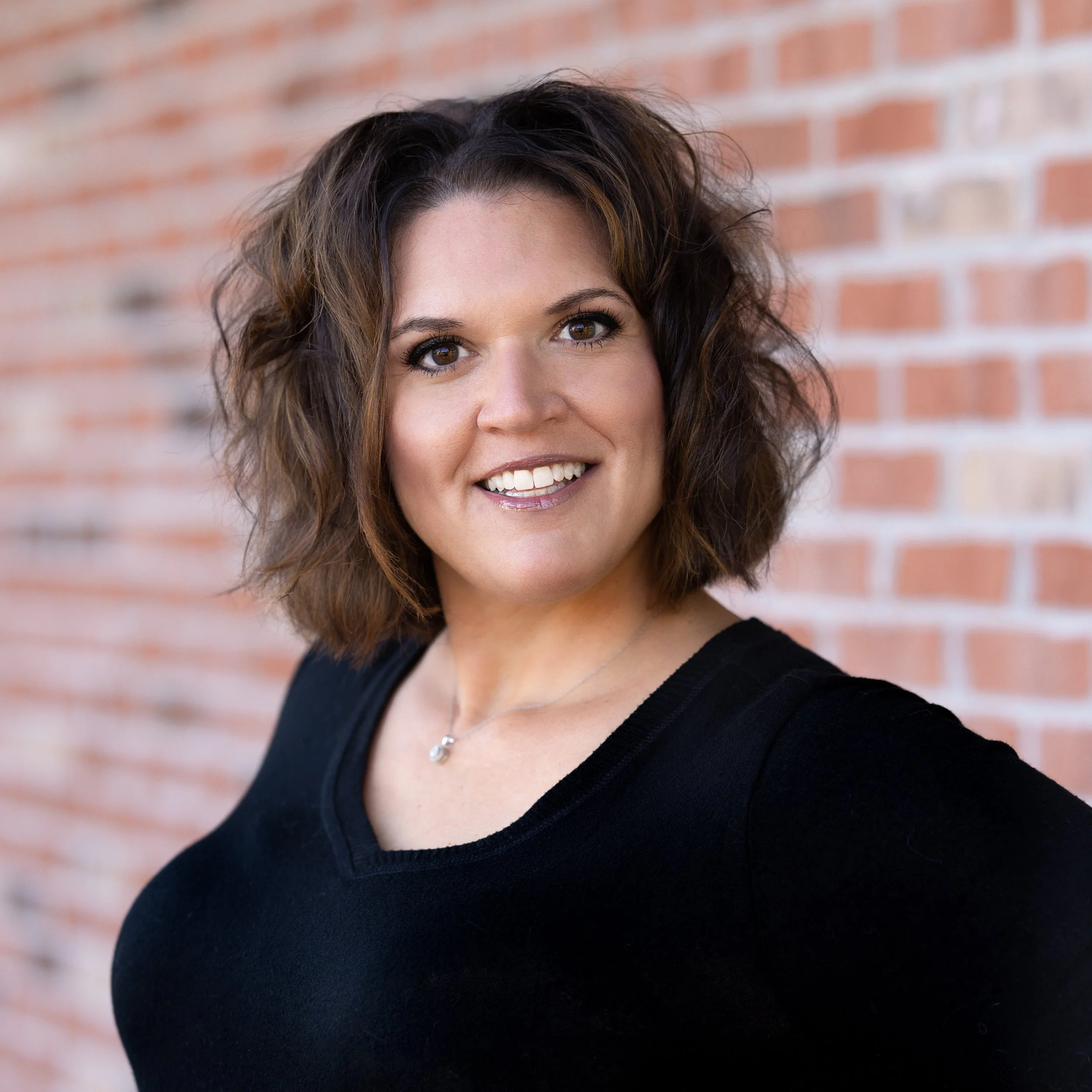 A woman with shoulder-length curly brown hair, smiling, wearing a black top, standing in front of a brick wall.