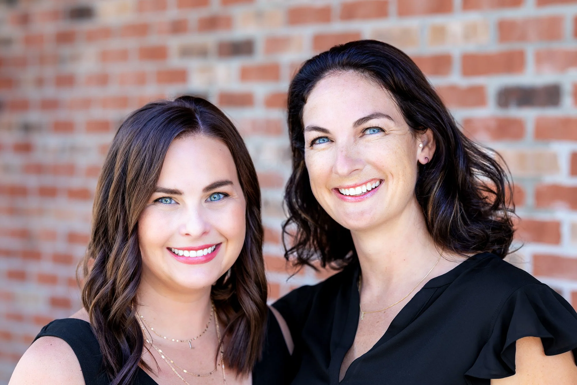 Two women smiling and standing in front of a brick wall, they are dentists at Beyond Your Smile Dentistry in Omaha