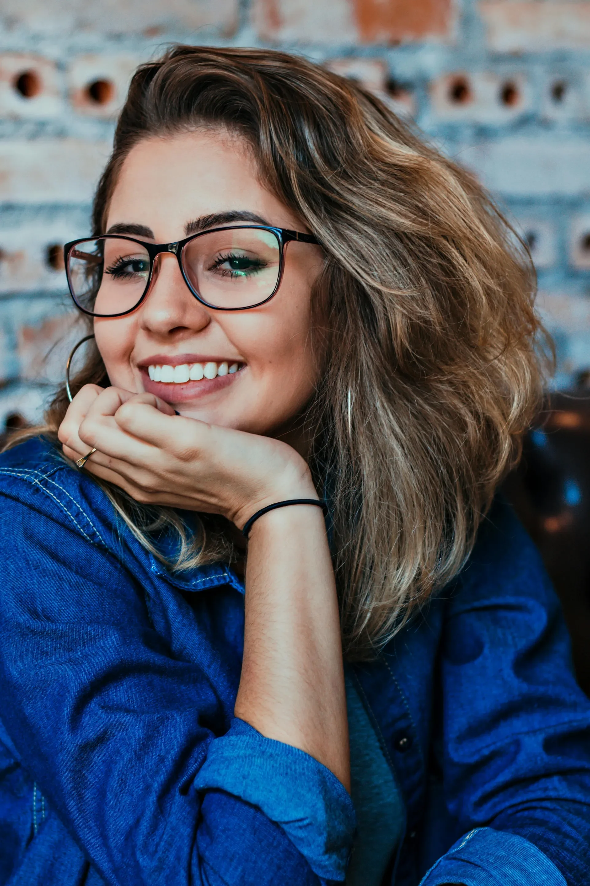 A young woman with glasses, curly hair, and hoop earrings, smiling and resting her chin on her hand, wearing a blue denim jacket, against a brick wall background.