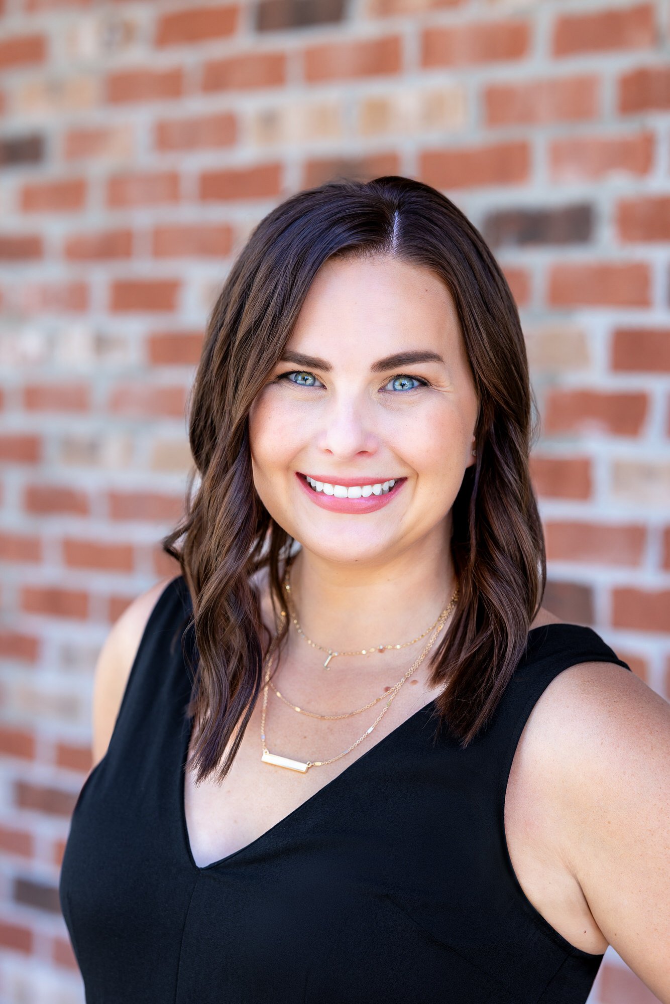 A staff member at Beyond Your Smile Dentistry, a woman with brown hair and blue eyes, smiling, wearing a black sleeveless top and layered gold necklaces, standing in front of a brick wall.