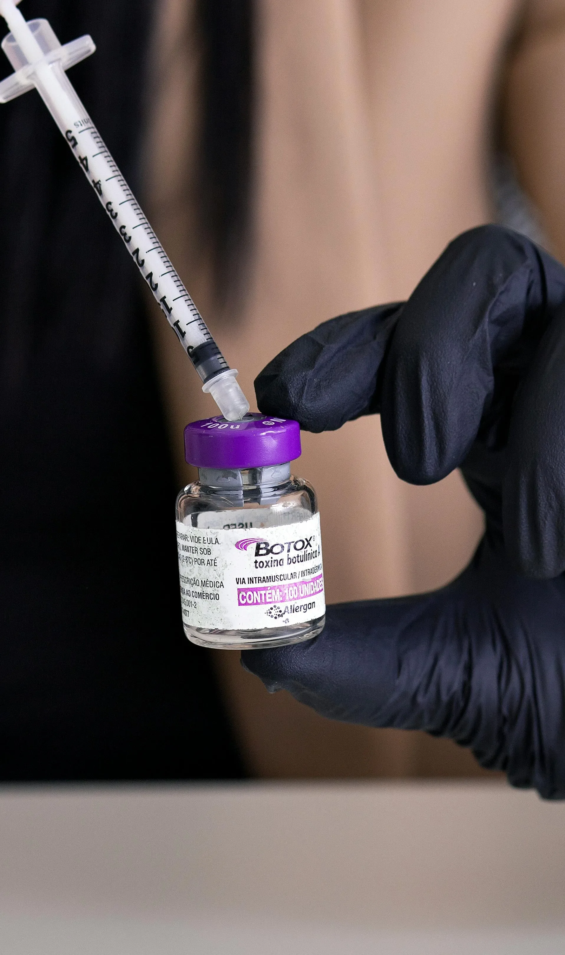 A person wearing black gloves holding a small vial labeled 'botox' with a syringe injecting into it.