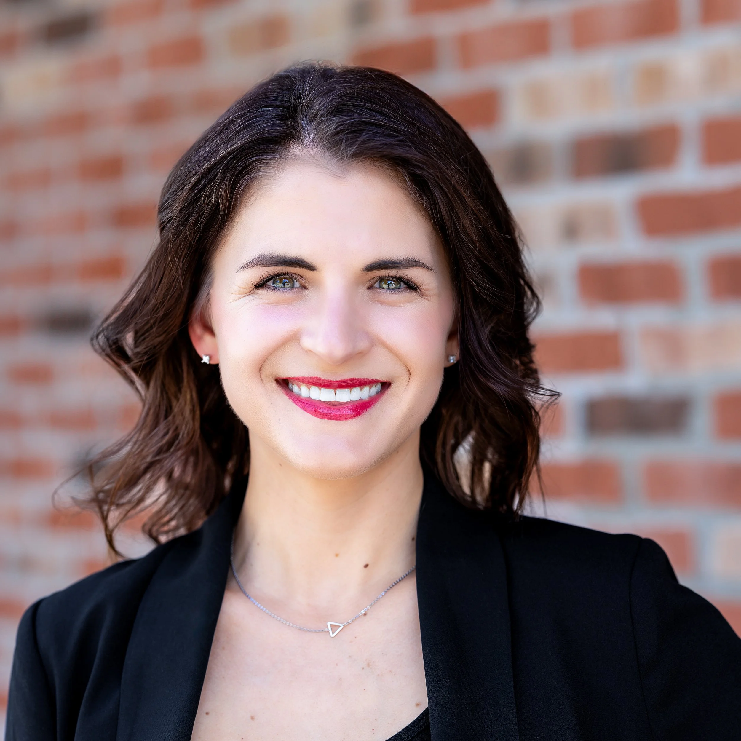 A woman with shoulder-length dark brown hair, wearing a black top and blazer, smiling in front of a red brick wall.