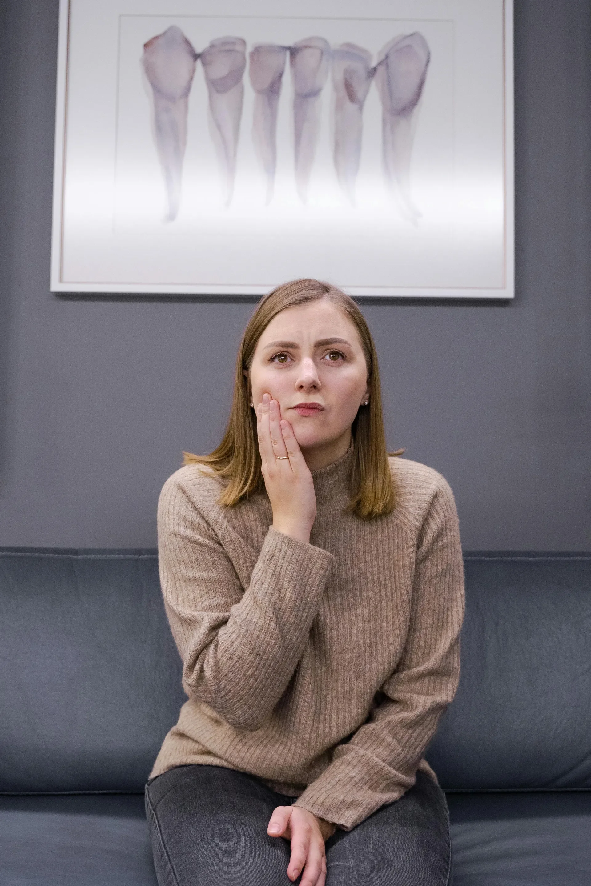 A woman with shoulder-length light brown hair, wearing a brown sweater, sits on a black couch in front of a gray wall with a framed picture of human teeth or jaws.