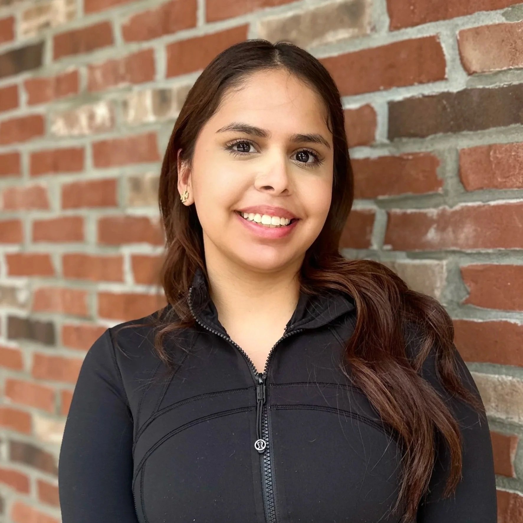 A young woman with long brown hair, smiling, wearing blue scrubs, standing in front of a red brick wall.