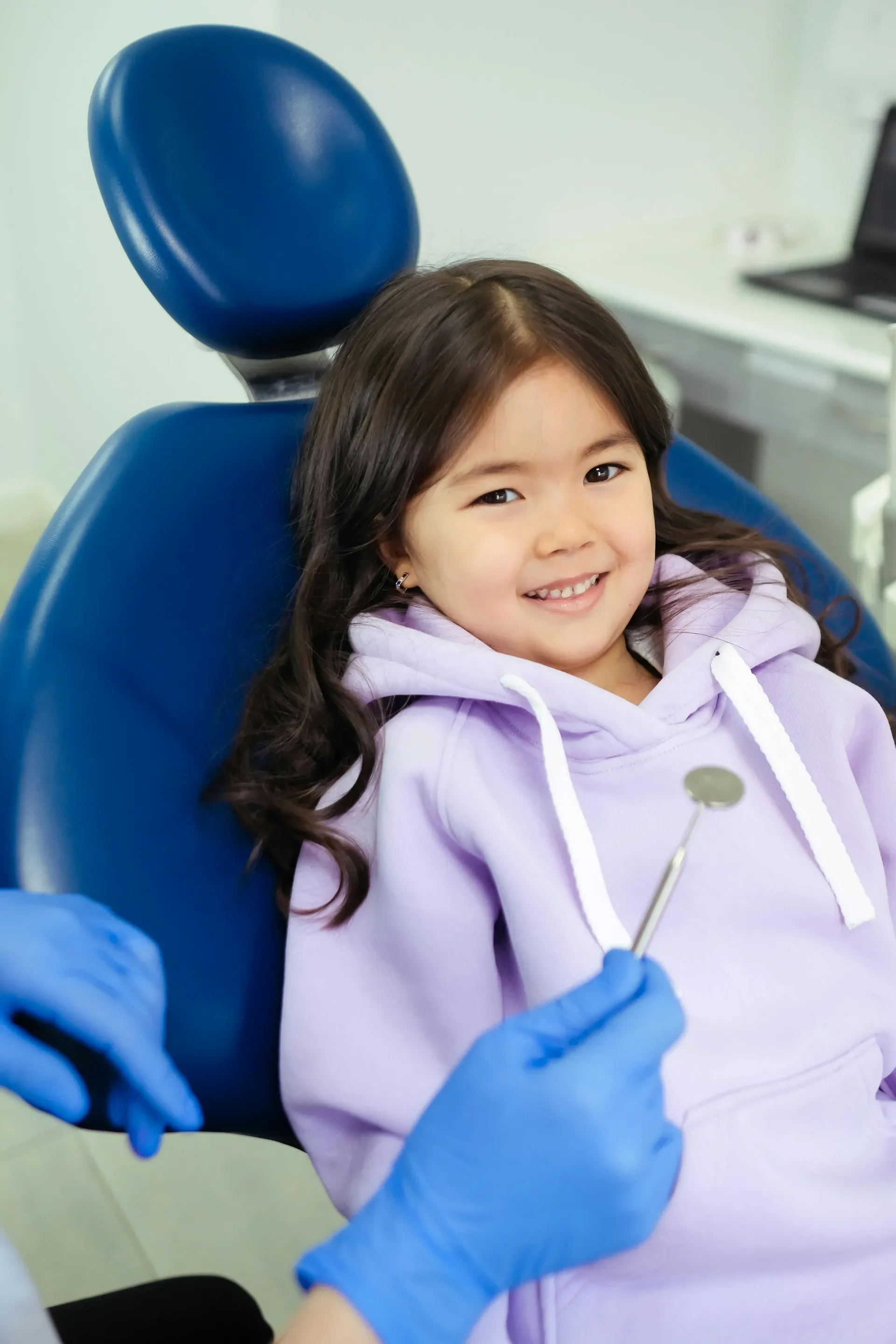 A young girl sitting in a dental chair, smiling at the camera, while a dental professional prepares to take a dental impression with a dental mirror.