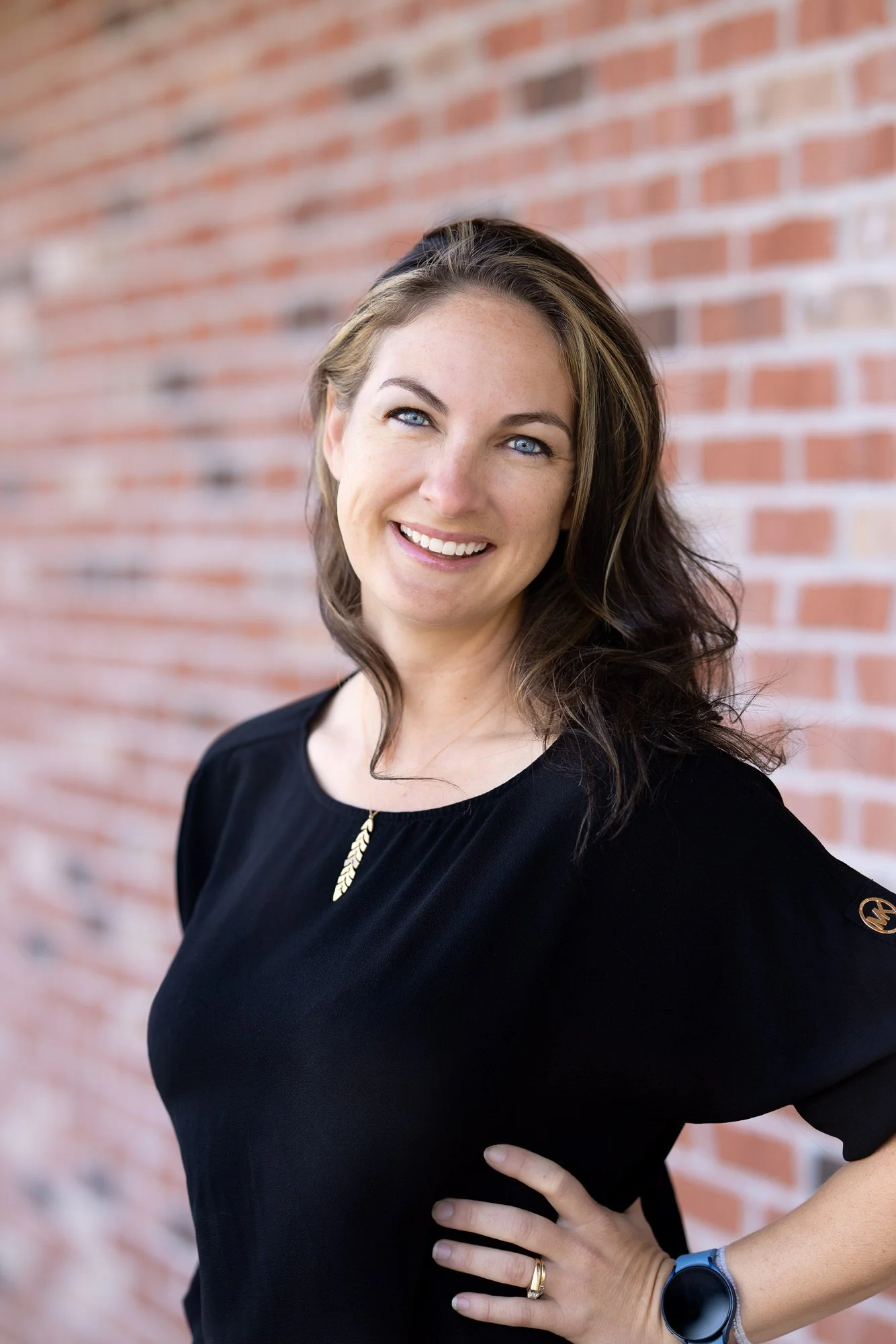 A staff member at Beyond Your Smile Dentistry, a woman with shoulder-length brown hair and blue eyes smiling, standing against a red brick wall, wearing a black top, a gold necklace, and a smartwatch.