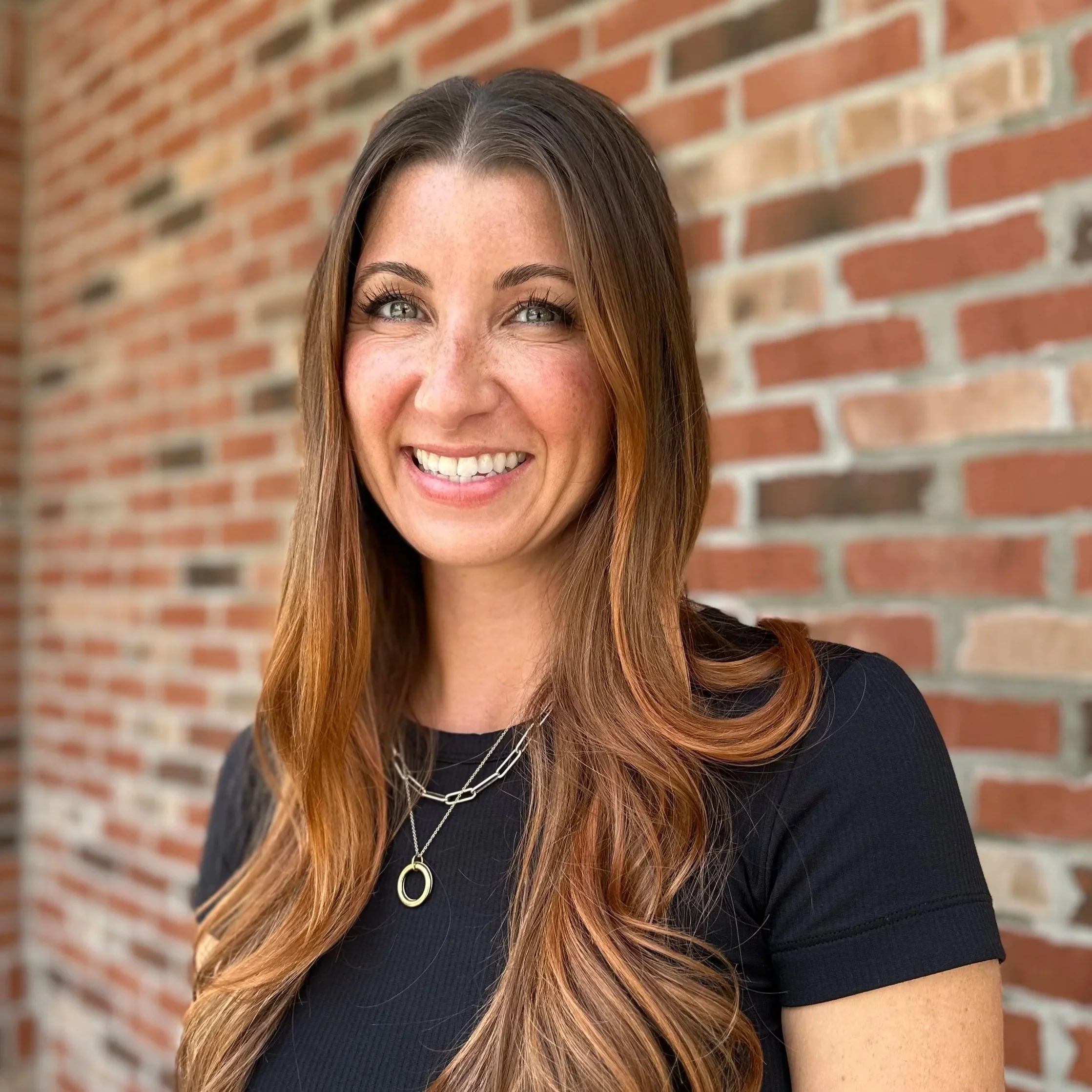 A woman with long wavy blonde hair wearing a black turtleneck sweater standing in front of a red brick wall.