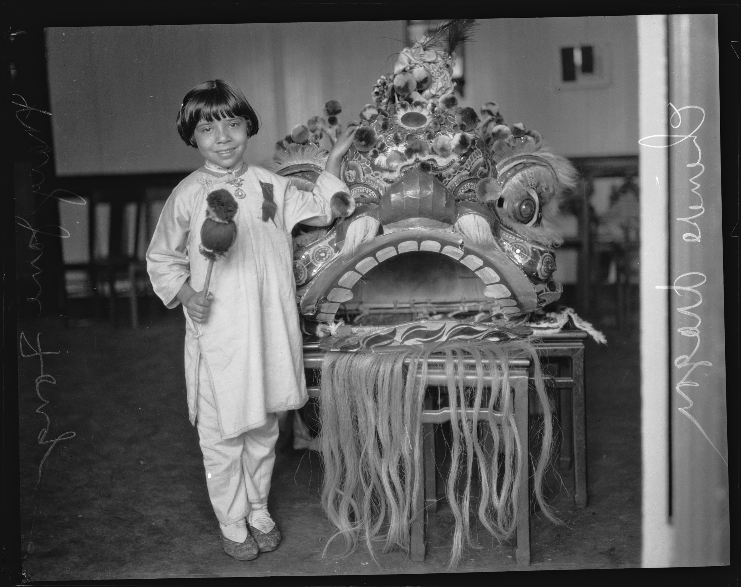 Mary Jane Fong holding a gong stick and standing next to a Chinese lion in Chinatown, Los Angeles. Photo via Los Angeles Times (1927)
