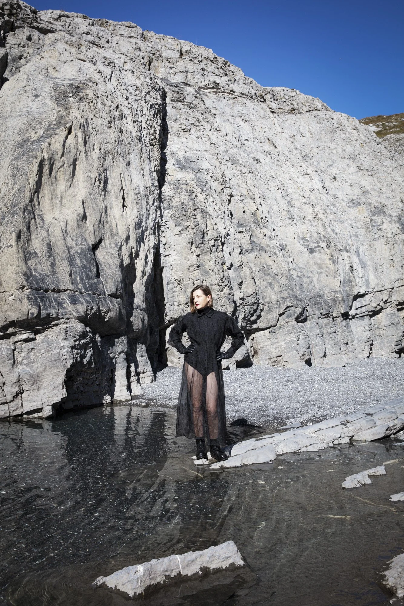 Une femme debout sur une roche dans une rivière, portant une robe noire en dentelle transparente, avec une grande falaise rocheuse en arrière-plan et un ciel bleu clair.