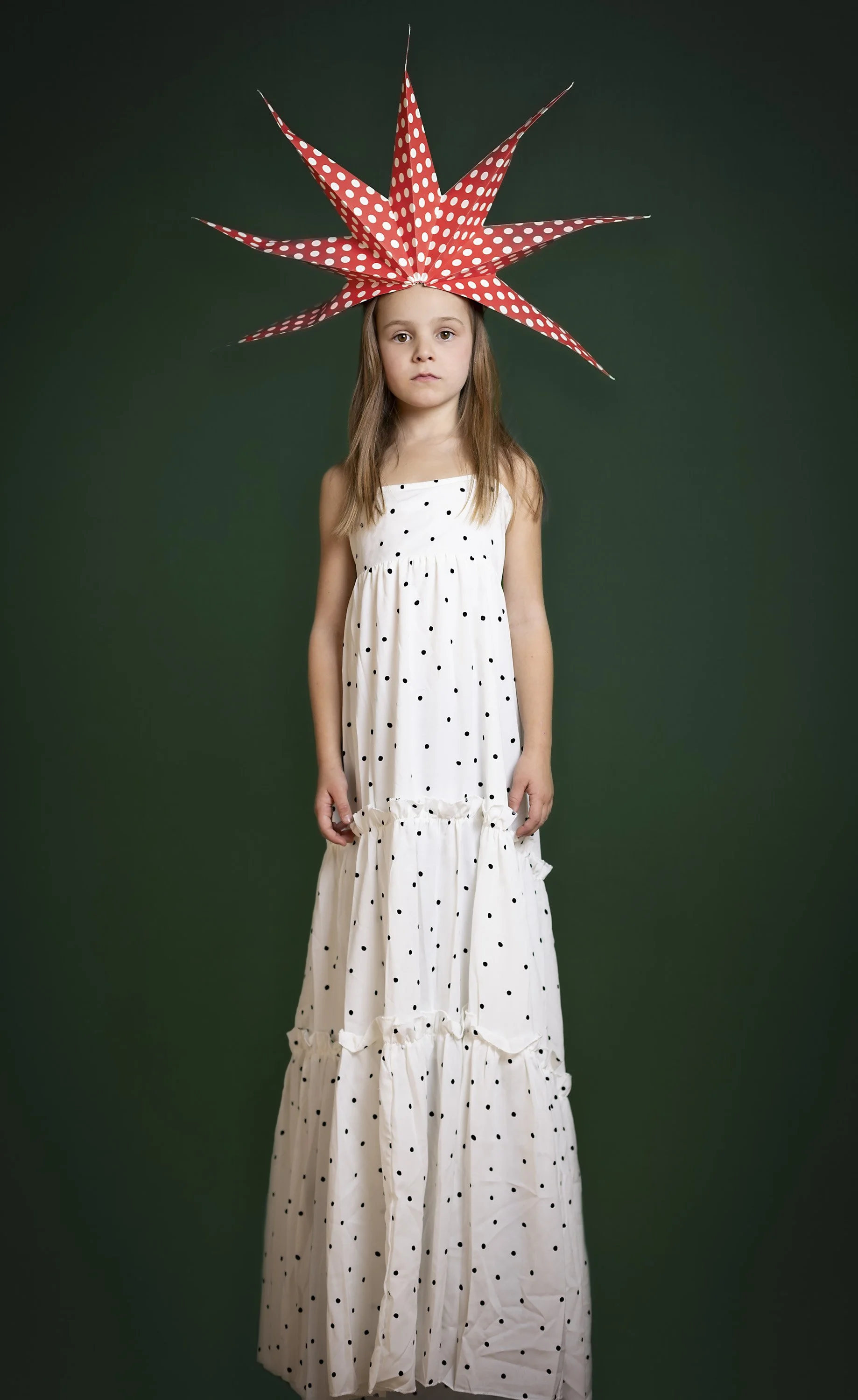Une jeune fille porte un chapeau rouge à pois blancs, ressemblant à une étoile ou un soleil, et une robe blanche à pois noirs, se tenant contre un fond vert.
