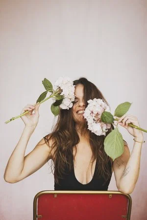 Femme souriante tenant des fleurs blanches, cachant partiellement son visage, contre un fond neutre.