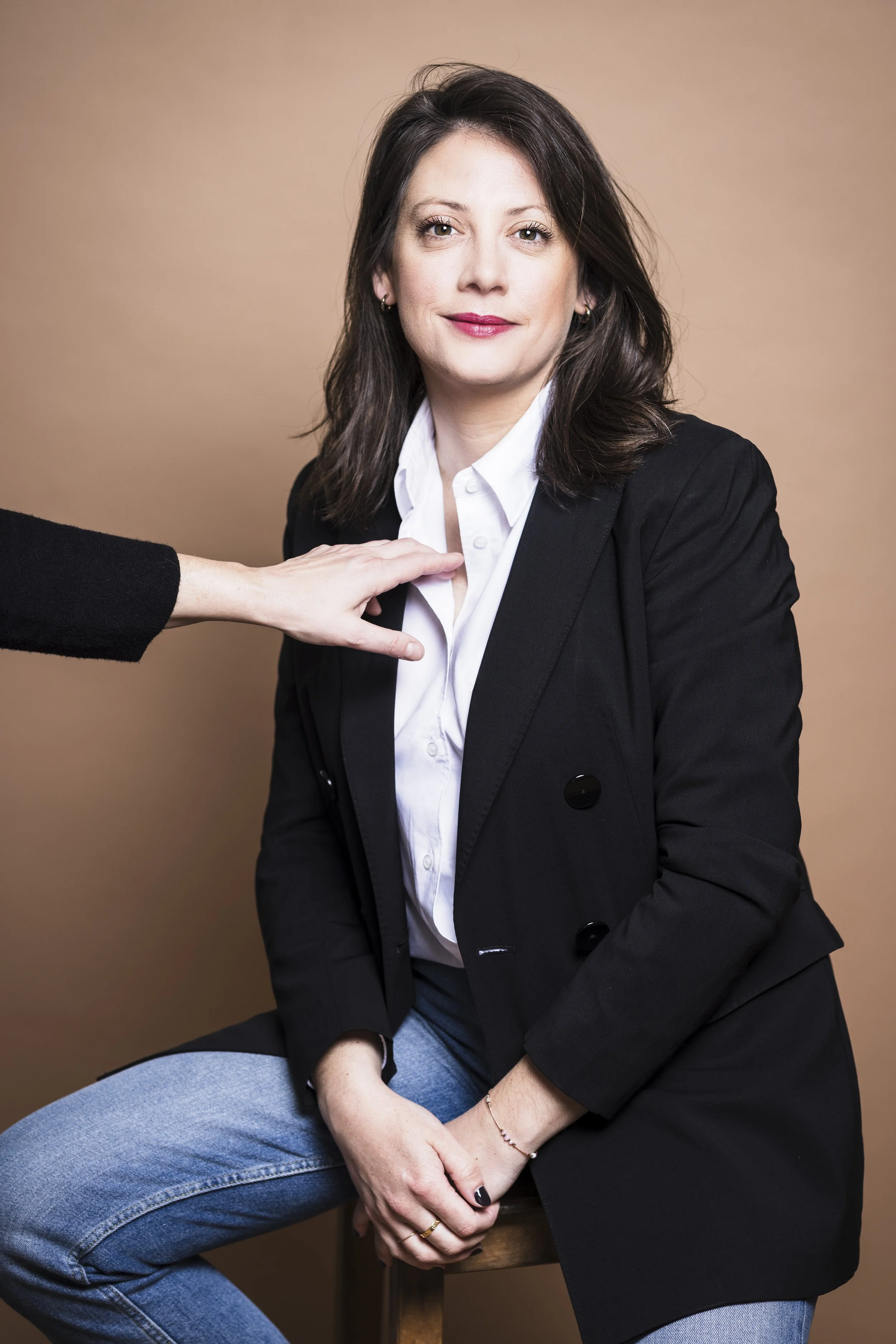 Une femme assise portant une veste noire et une chemise blanche, avec une main d'une autre personne posée sur sa poitrine, devant un fond beige.