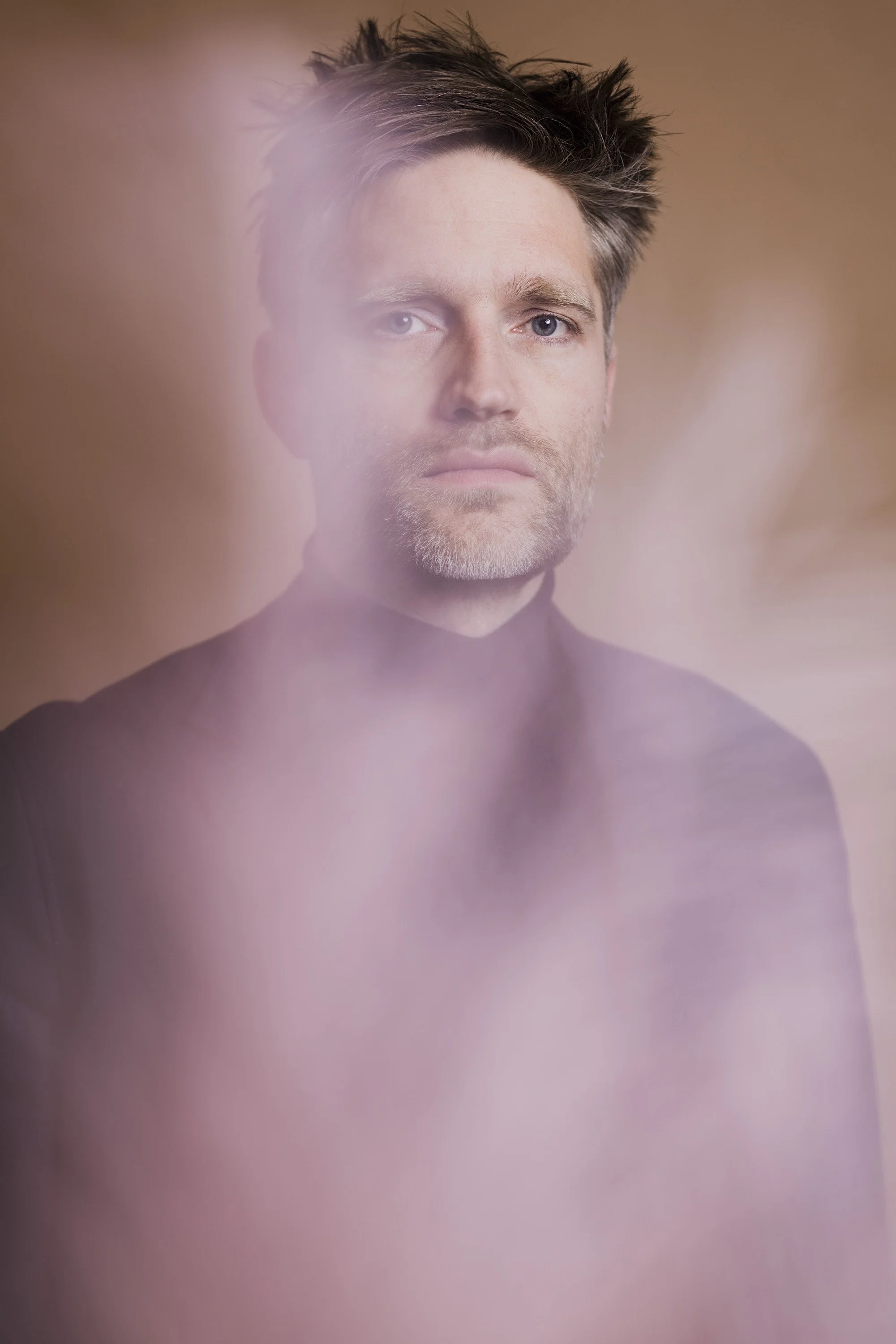 Un portrait d'un homme avec des cheveux bruns et une barbe, portant un t-shirt noir, avec une brume rose-violet en premier plan, floue.