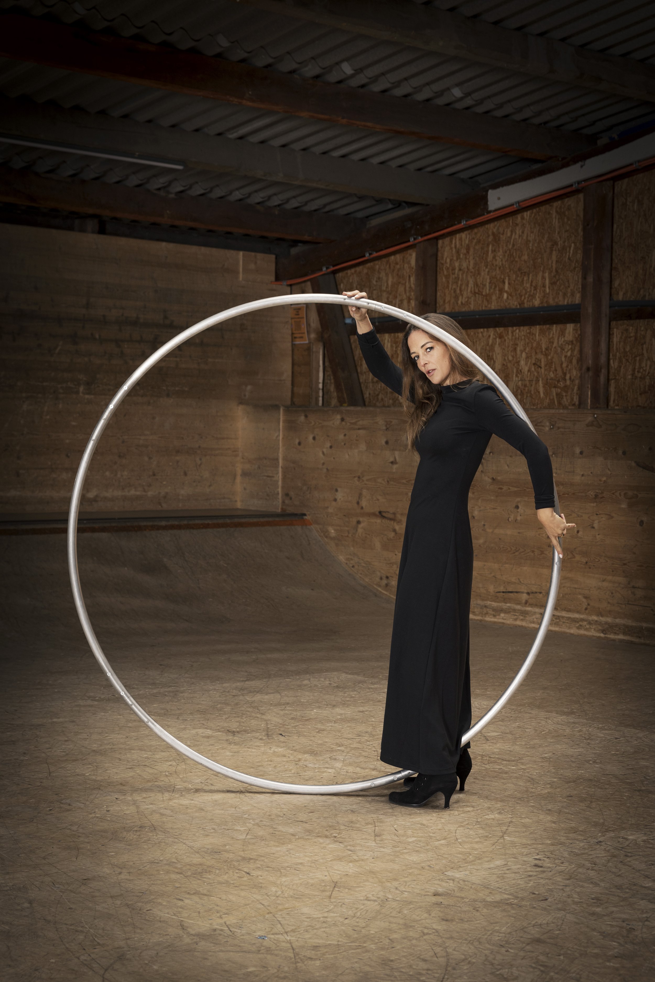 Femme en robe noire et talons, tenant un grand cerceau en métal dans un lieu en bois, probablement un skatepark ou un espace de pratique acrobatique.