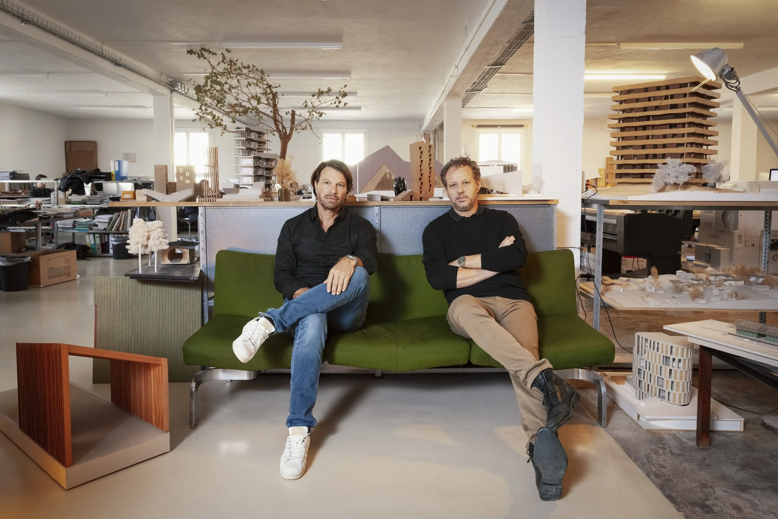 Deux hommes assis sur un canapé vert dans un atelier de design ou d'architecture rempli de maquettes, plans et matériaux de modélisme.