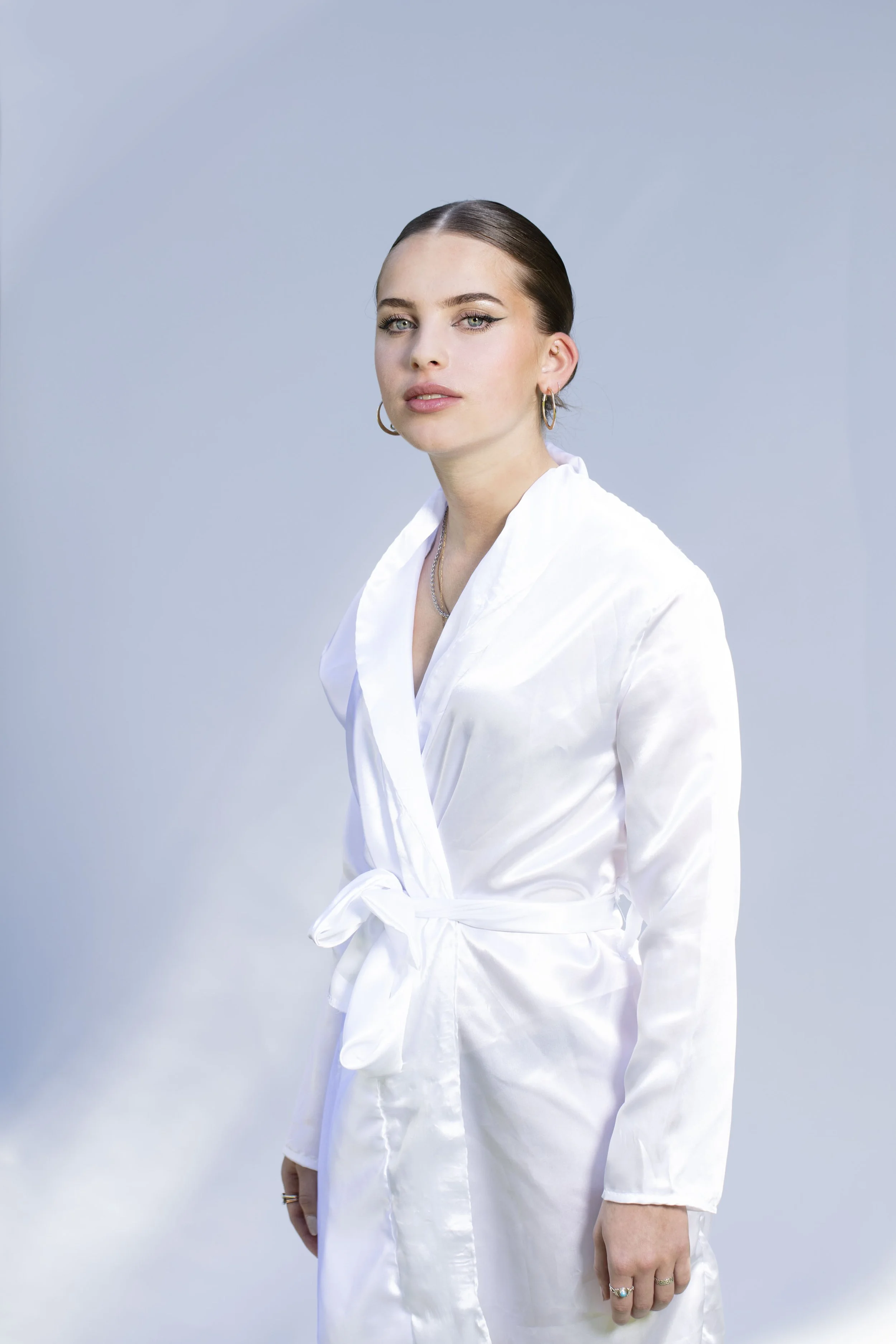 Une femme portant une robe de chambre blanche en satin, cheveux bruns tirés en arrière, portant des boucles d'oreilles et des colliers, posant contre un fond gris clair.