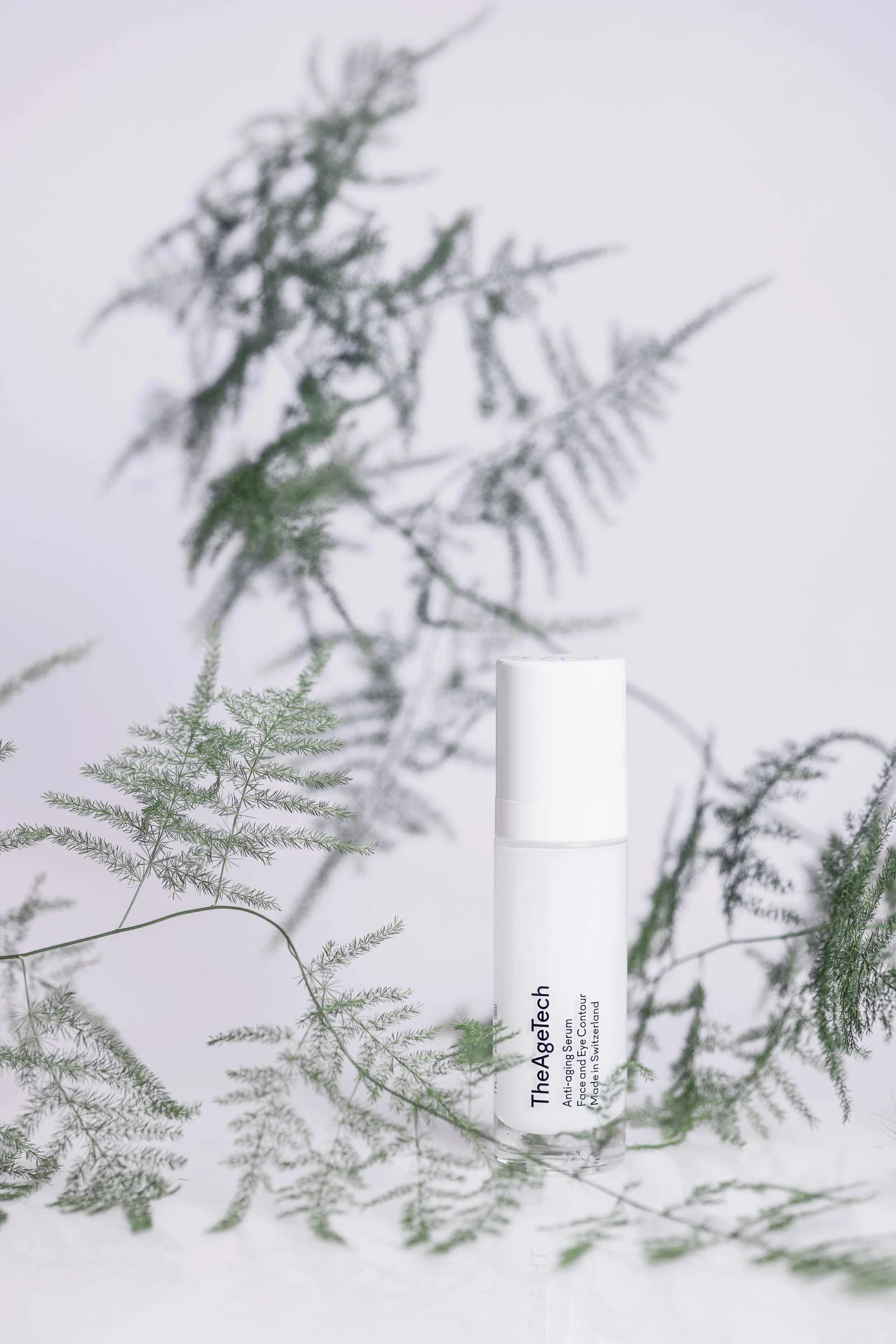 Un flacon blanc de sérum anti-âge TheAGetaTech entouré de branches de fougère vertes, sur fond blanc.