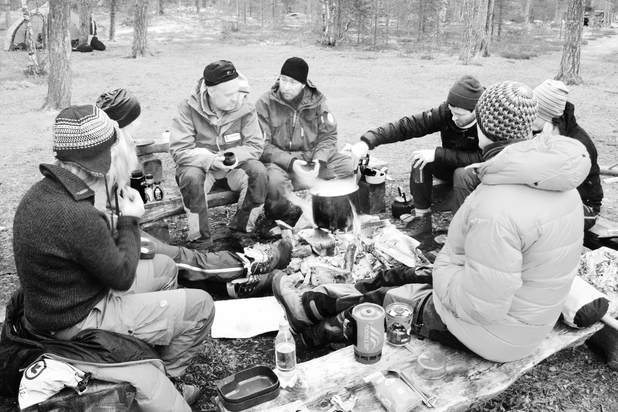 En gruppe sitter sammen rundt et bål i skogen, fotografert i svart-hvitt.