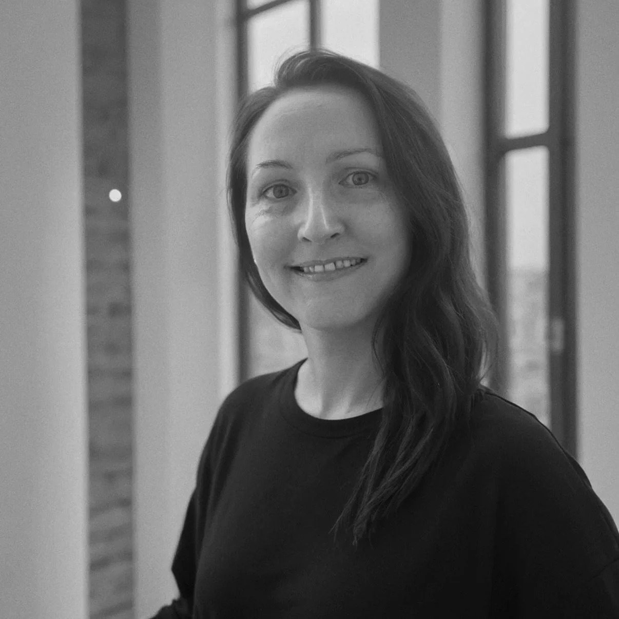 Black and white photo of a smiling woman with long hair, wearing a dark shirt, in front of windows with a brick wall outside.