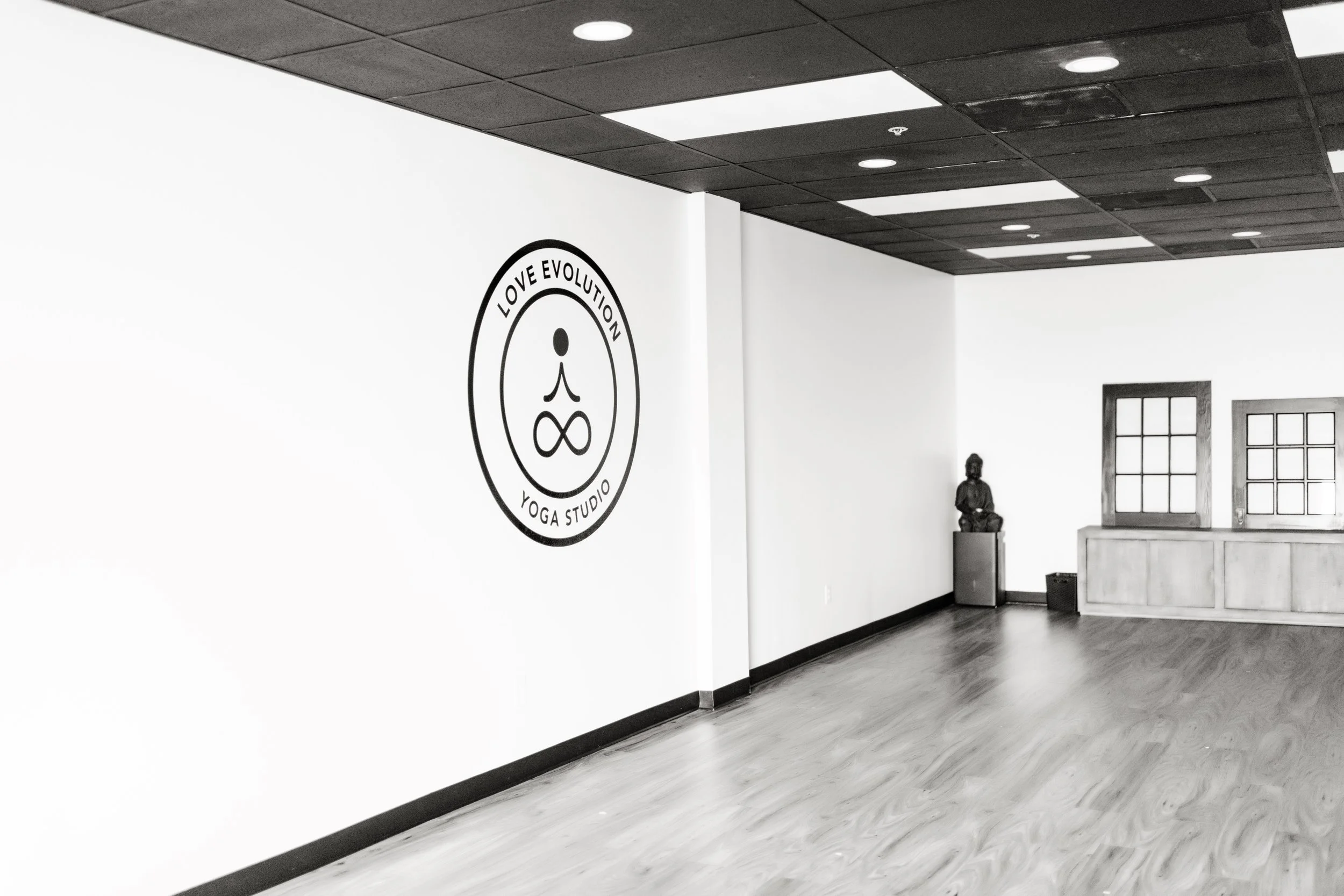 Interior of a yoga studio with wooden flooring, white walls, and a black ceiling. There is a logo on the wall that reads "Love Evolution Yoga Studio" with a stylized face and infinity symbol. A statue of a seated figure is placed on a pedestal near two windows, which have wooden frames and multiple panes.
