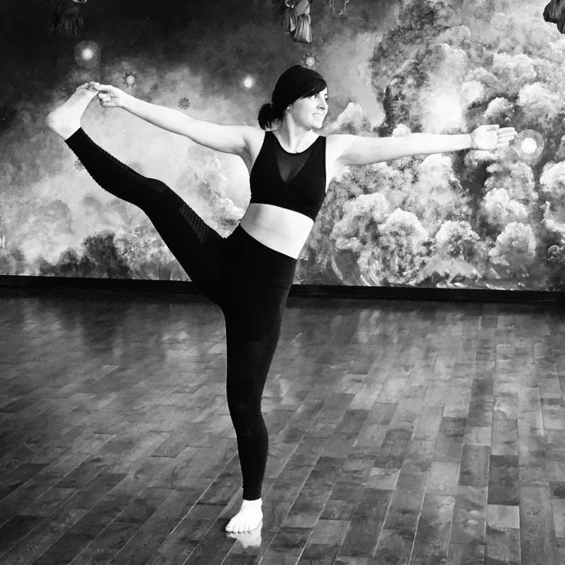 A woman with dark hair and headband in black leggings and a black tank top doing standing hand to big toe with side extension..