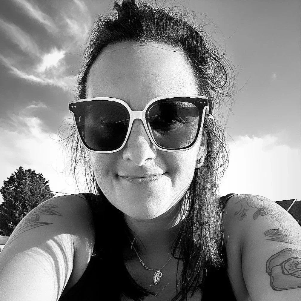 A woman with dark hair, wearing large sunglasses, smiling, outside with trees and a cloudy sky in the background.