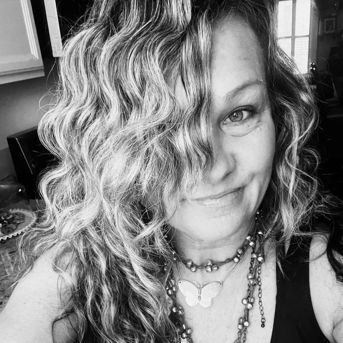 A woman with curly hair, smiling slightly, wearing layered necklaces, including a butterfly pendant, in a room with a window in the background.
