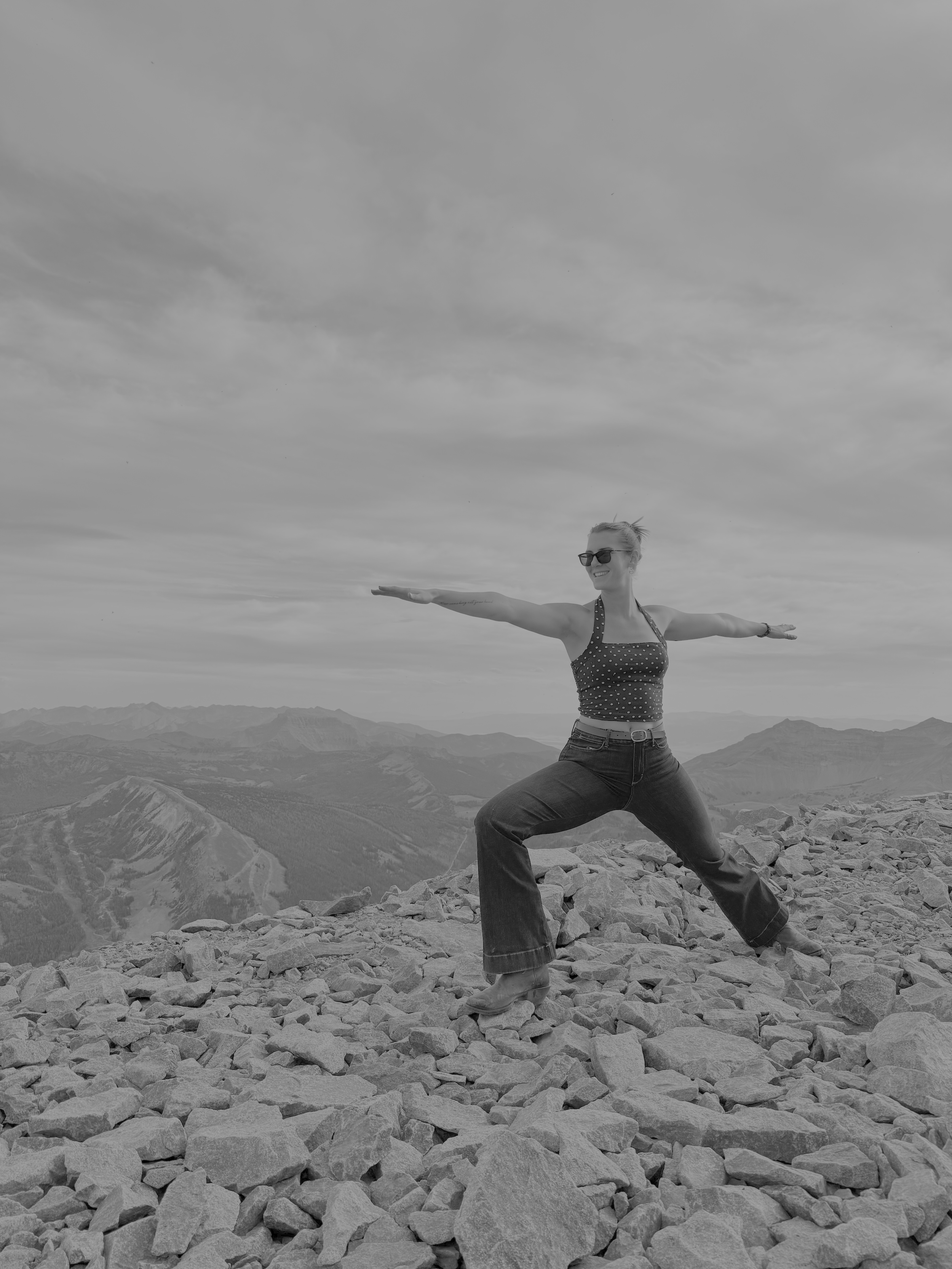 A woman doing warrior two yoga pose on mountain top in jeans and cowboy boots.