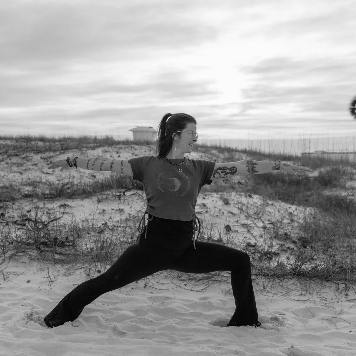 A woman with curly hair, smiling slightly, wearing layered necklaces, including a butterfly pendant, in a room with a window in the background.