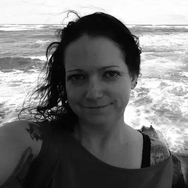 A woman with dark, wavy hair taking a selfie by the ocean with waves in the background, in black and white.