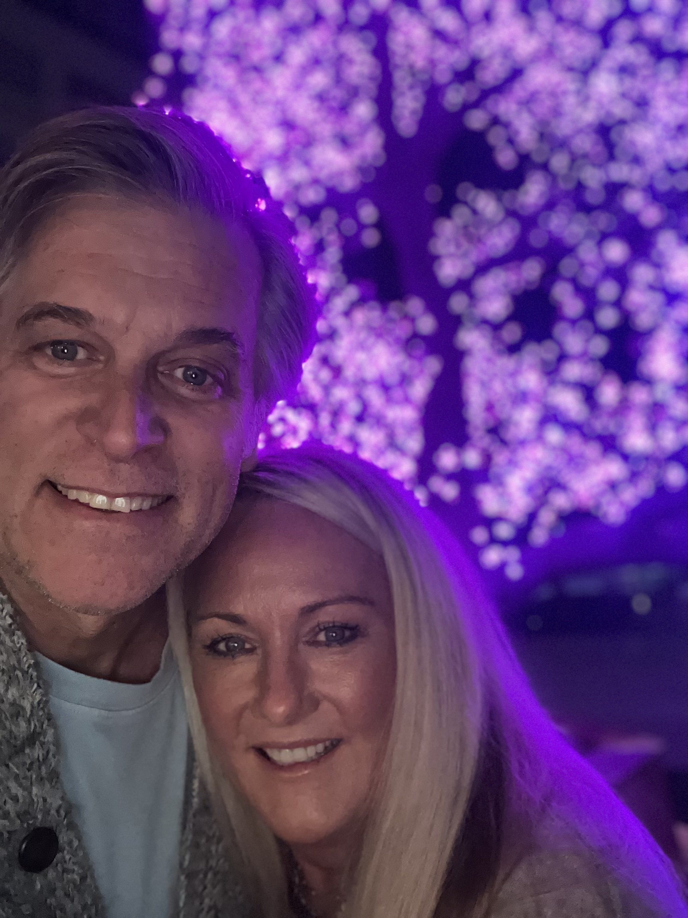 A smiling man and woman taking a selfie together at night with purple-lit trees in the background.